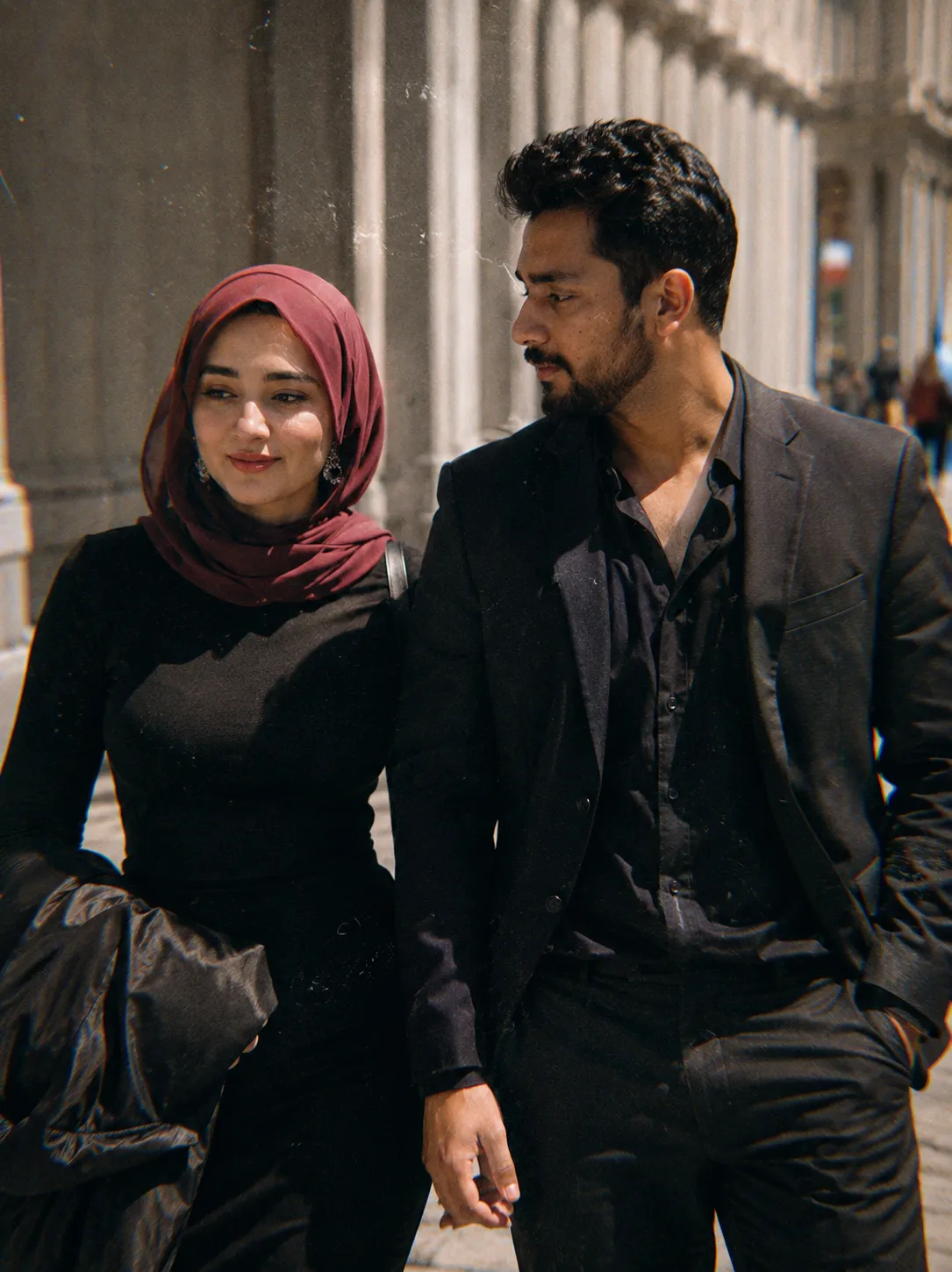 A cinematic candid medium shot portrait of a man and a woman walking side by side along a bright urban walkway, framed from the waist up.