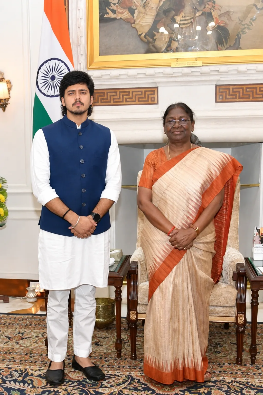 A formal, photorealistic portrait of a young Indian man standing respectfully beside a distinguished elderly Indian woman in an elegant