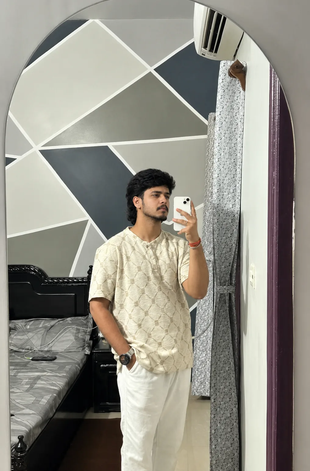 A highly realistic full-body mirror selfie of a young South Asian man standing casually in a modern bedroom, captured through a large