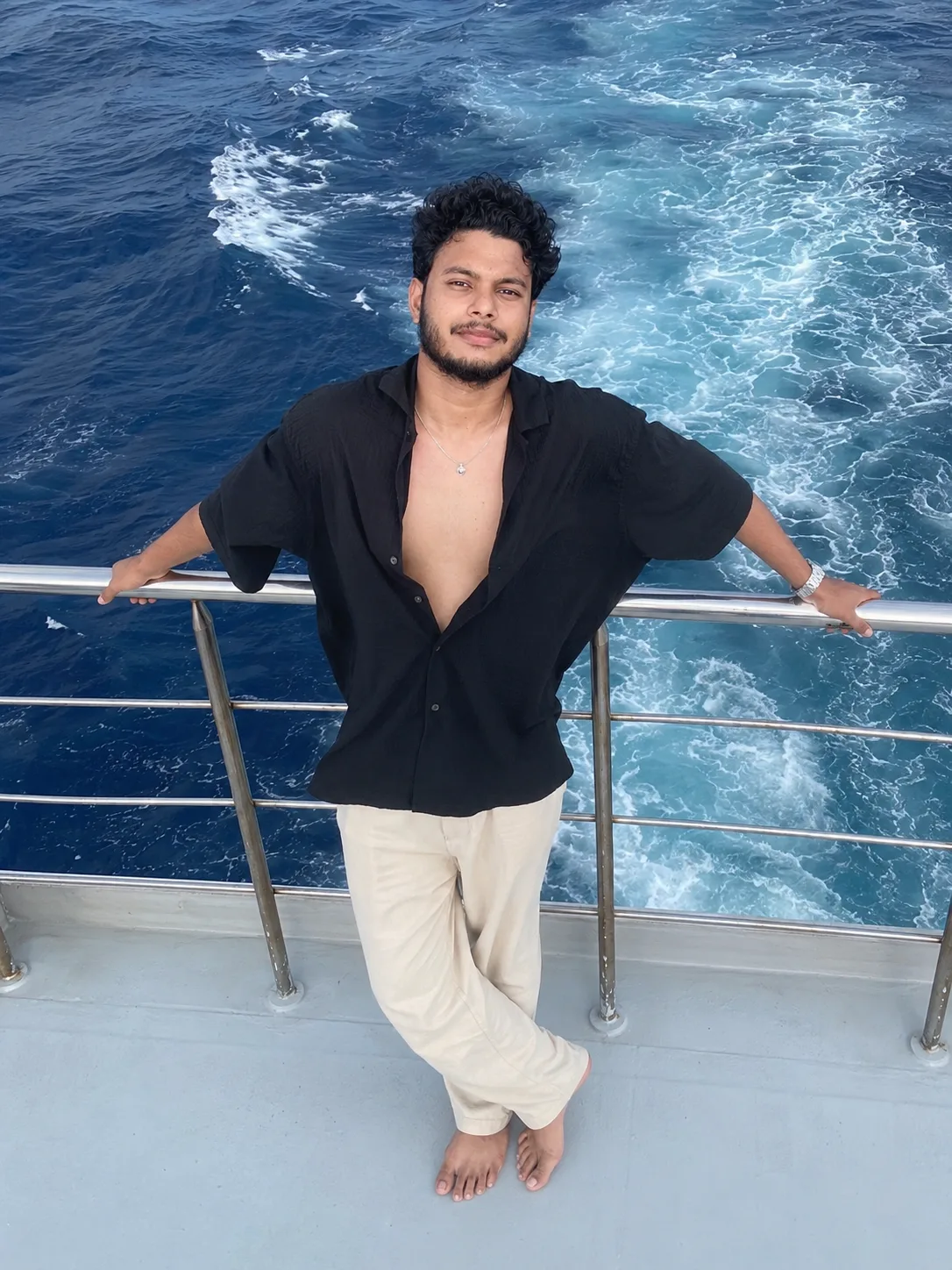 A young Indian man with curly black hair, well-groomed short beard and mustache, standing barefoot on the gray deck of a moving boat or