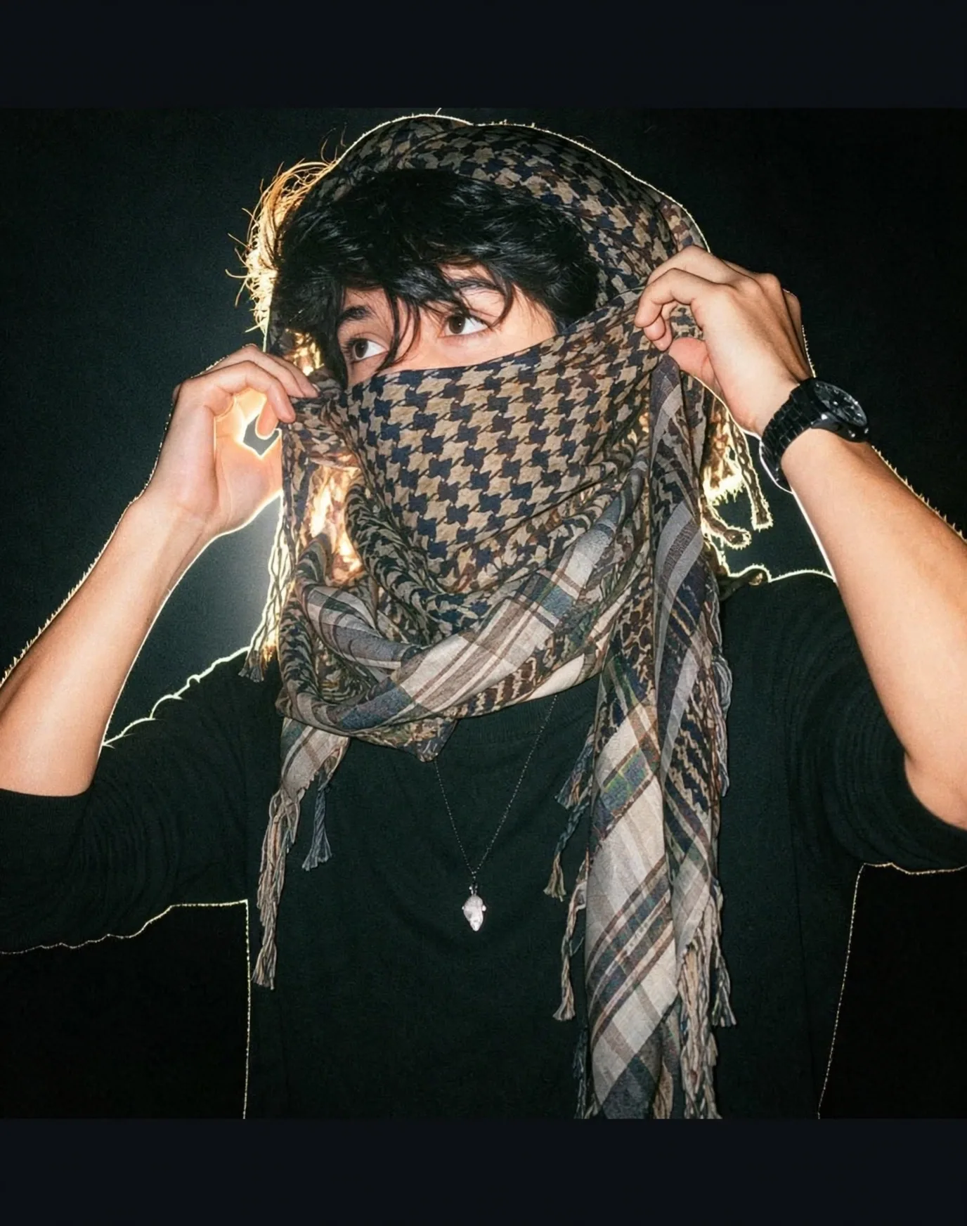 A young South Asian man with messy black hair, wearing a black long-sleeve t-shirt, a thin silver pendant necklace with a small white