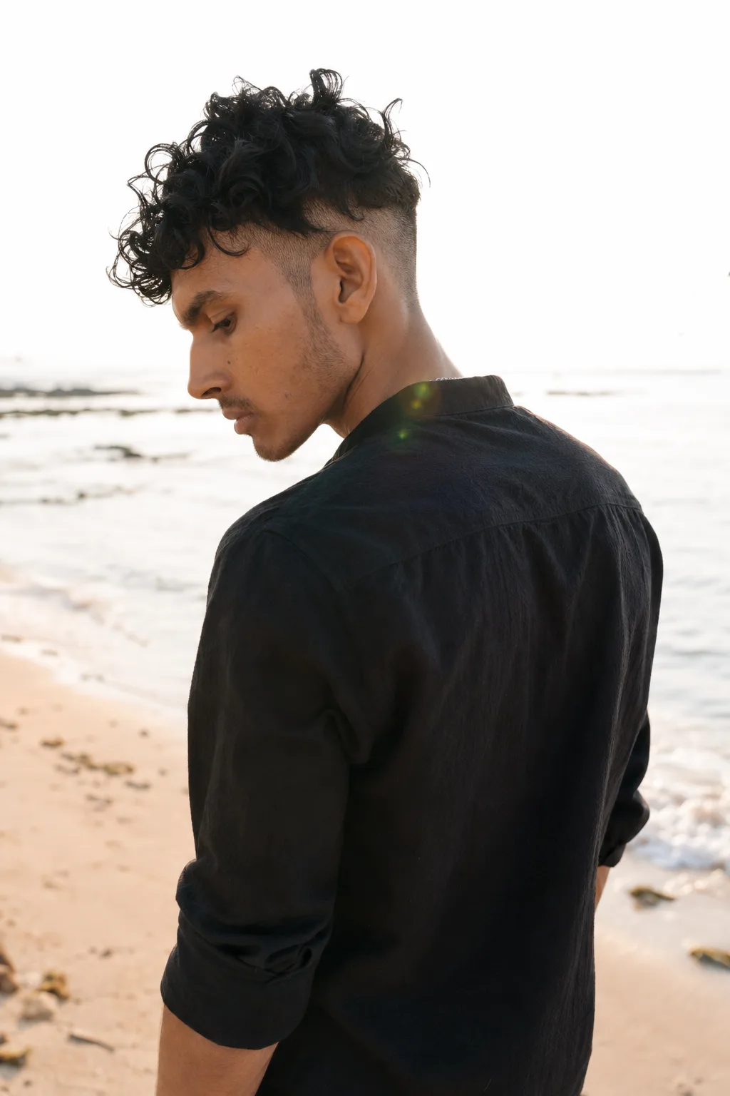 Candid high-fashion editorial portrait of a young man viewed from a three-quarters back angle, looking down.