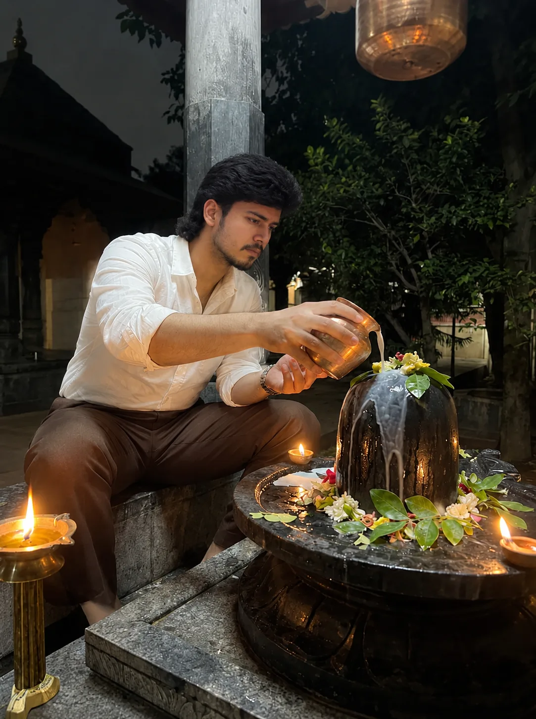 A highly realistic cinematic nighttime photograph of a young South Asian man sitting beside a sacred Shiva lingam inside a traditional