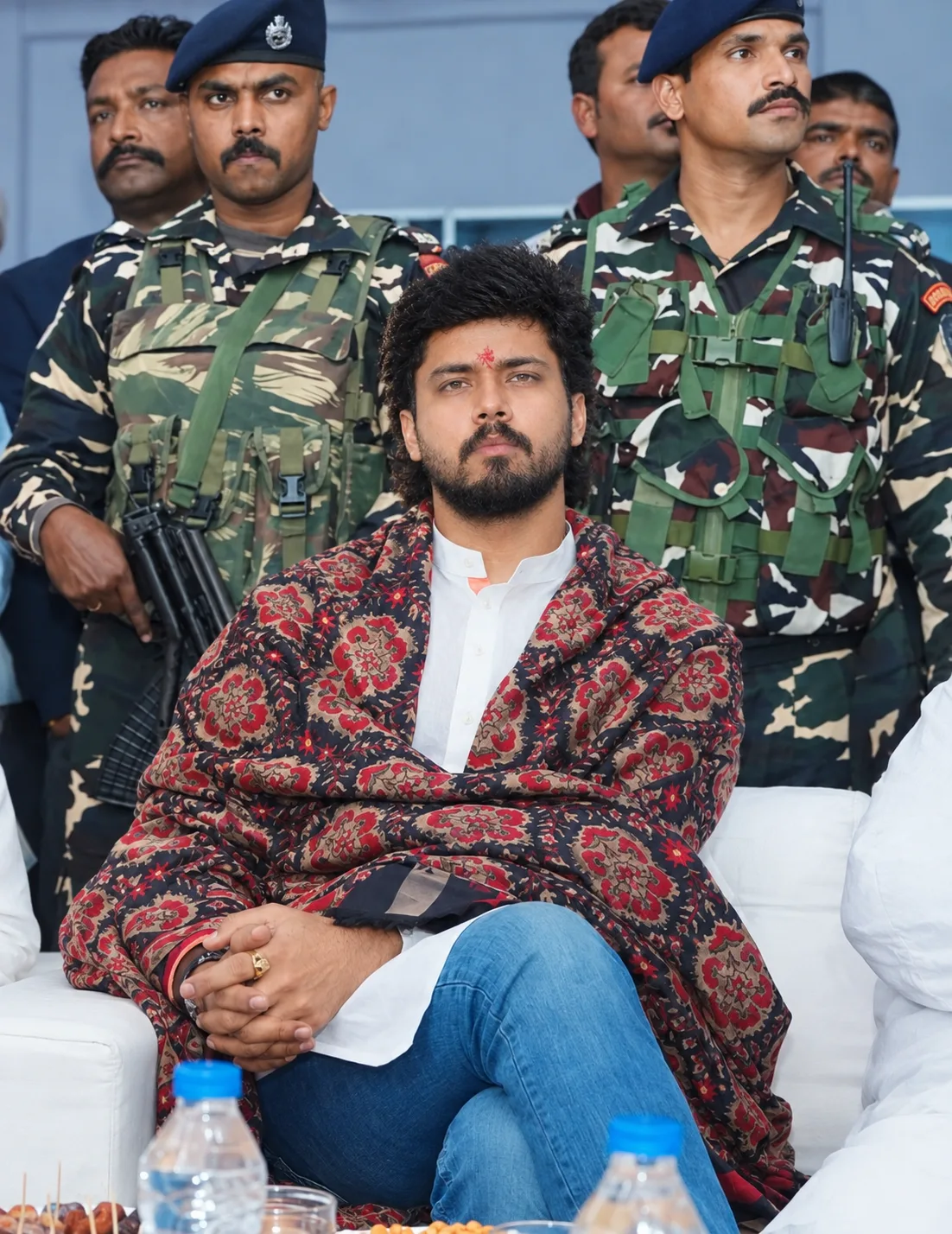 A photorealistic cinematic portrait of a young Indian man seated confidently at a formal outdoor event, surrounded by uniformed security