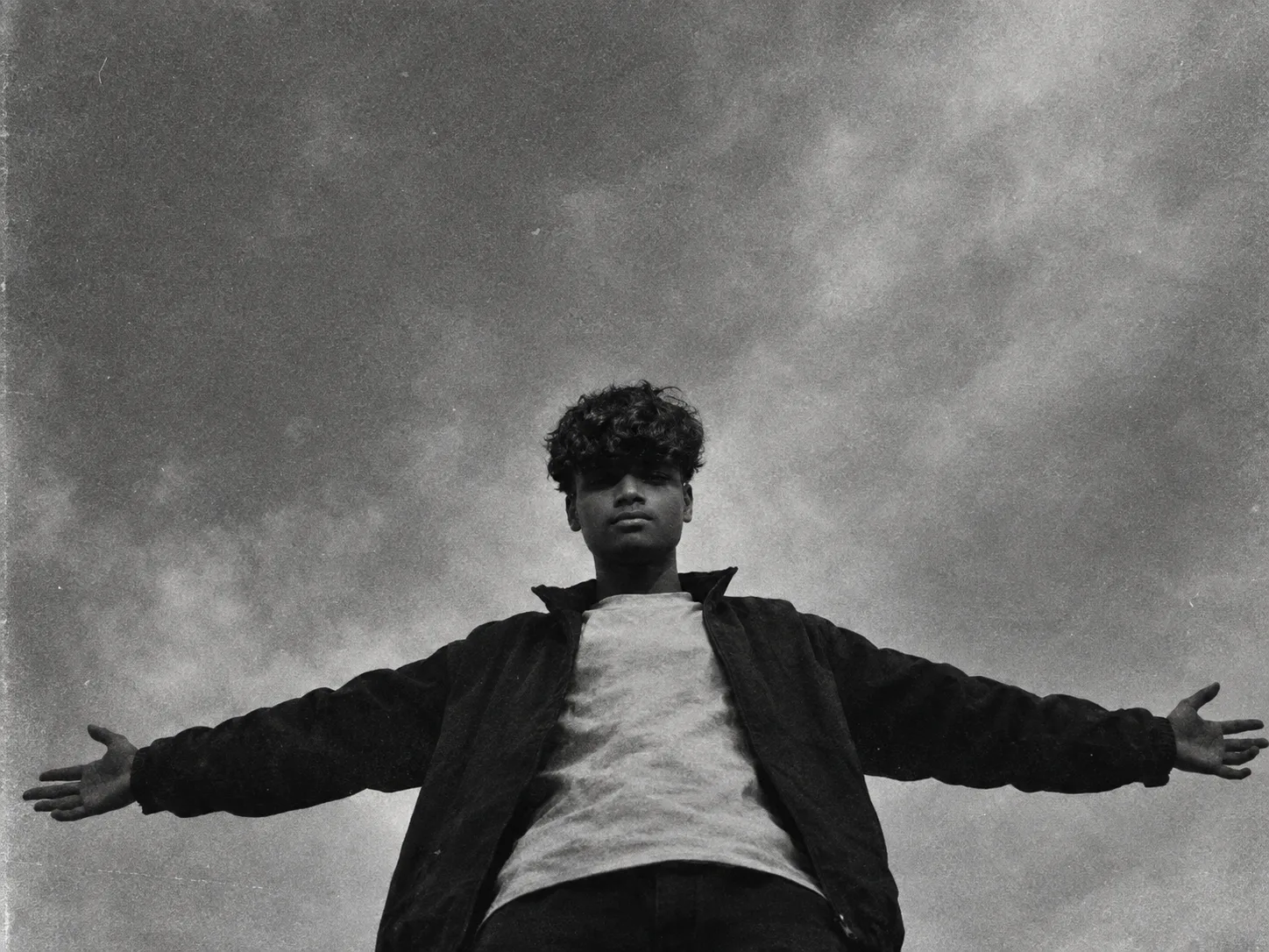 *A black-and-white grainy photo of a person standing with arms outstretched wide against a plain cloudy sky, viewed from a low angle,