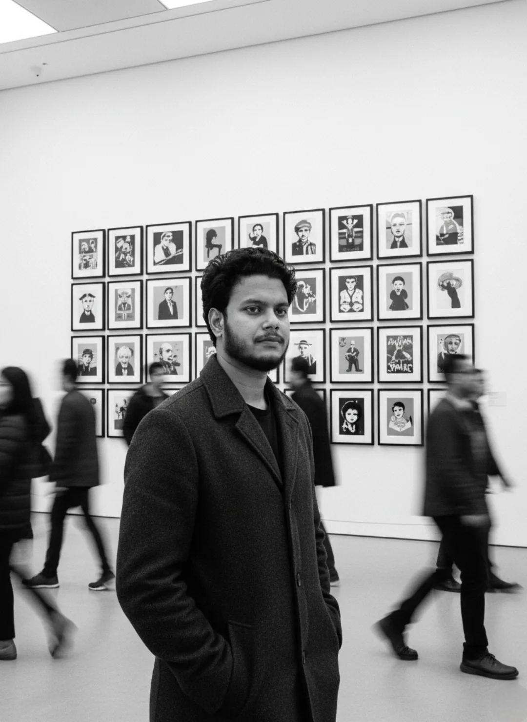 Black and white photograph of a young South Asian man with curly dark hair, short beard and mustache, standing confidently in the