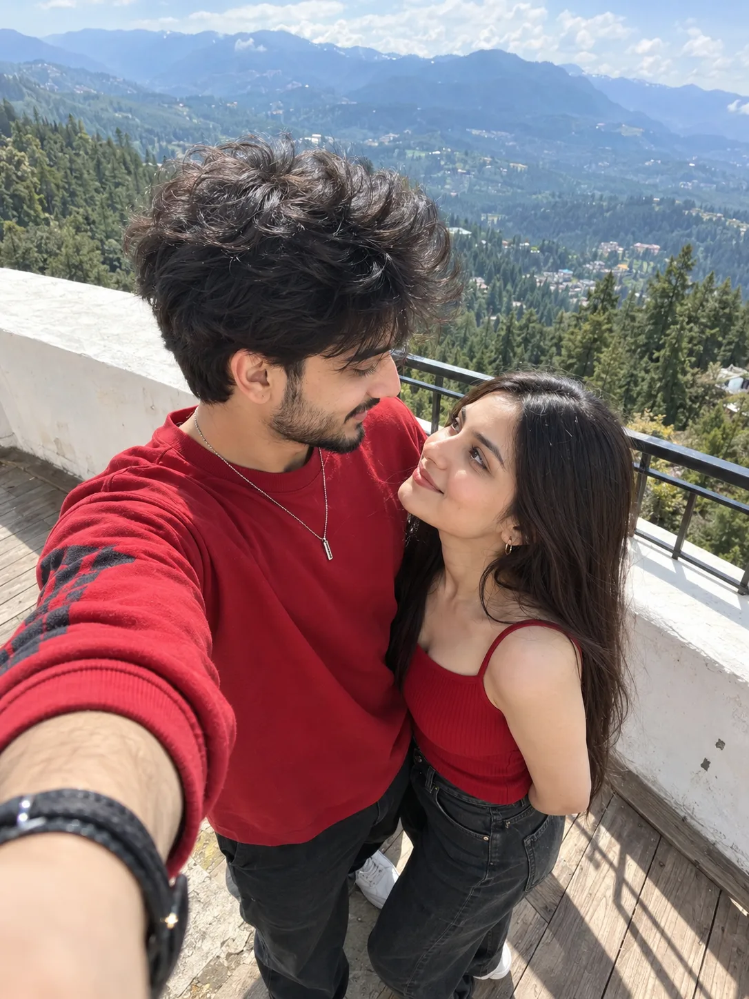 A candid smartphone front-camera selfie photograph capturing a young Indian couple on an outdoor balcony.