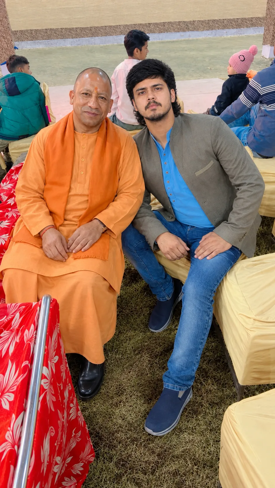 Create an ultra-realistic cinematic vertical portrait of two Indian men seated together at an outdoor public gathering, captured in a