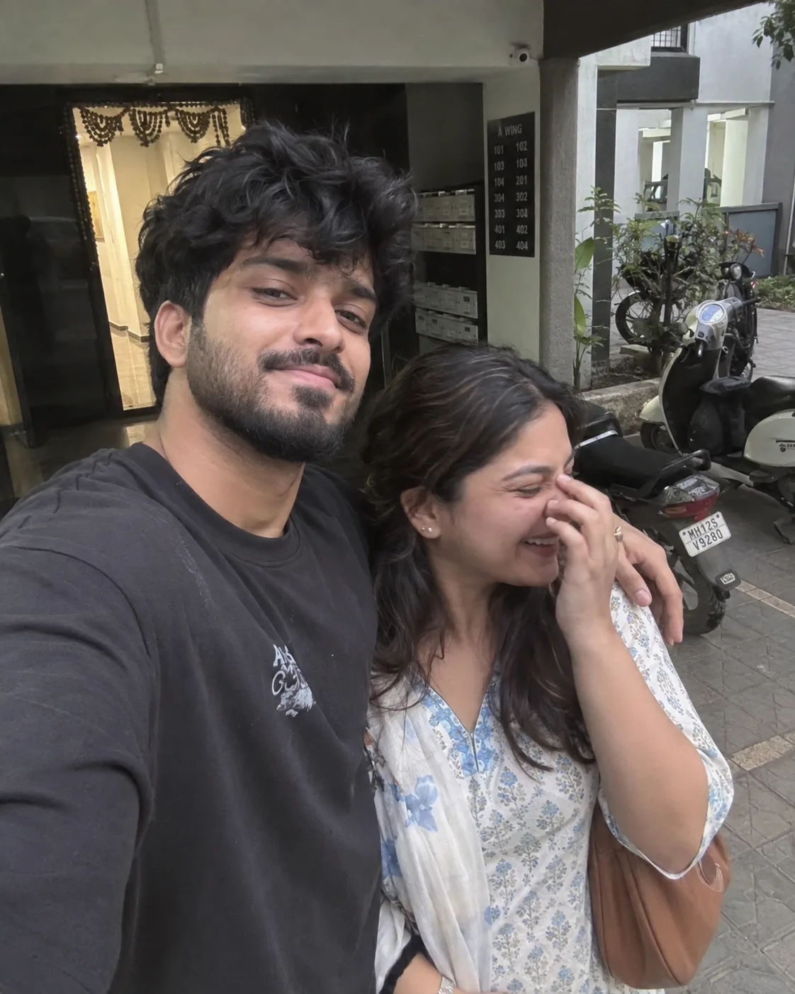 Photorealistic 4:5 vertical candid iPhone photograph of a young Indian couple standing together outside their apartment building.
