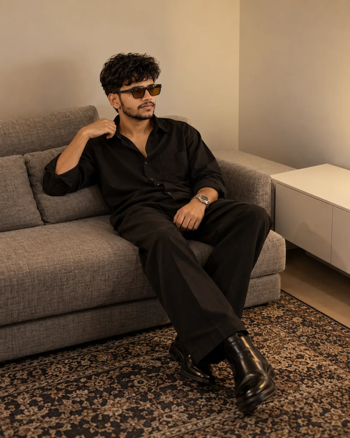 Ultra-realistic professional DSLR photograph of a young man seated casually on a modern gray fabric sofa in a minimalist warm-toned