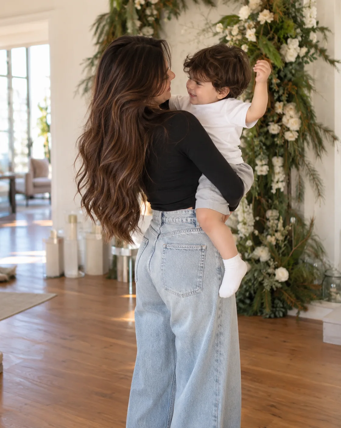 A HEARTWARMING INDOOR PHOTOGRAPH, FROM A THREE-QUARTER BACK ANGLE, OF A WOMAN WITH LONG, FLOWING BROWN WAVY HAIR HOLDING A TODDLER BOY IN