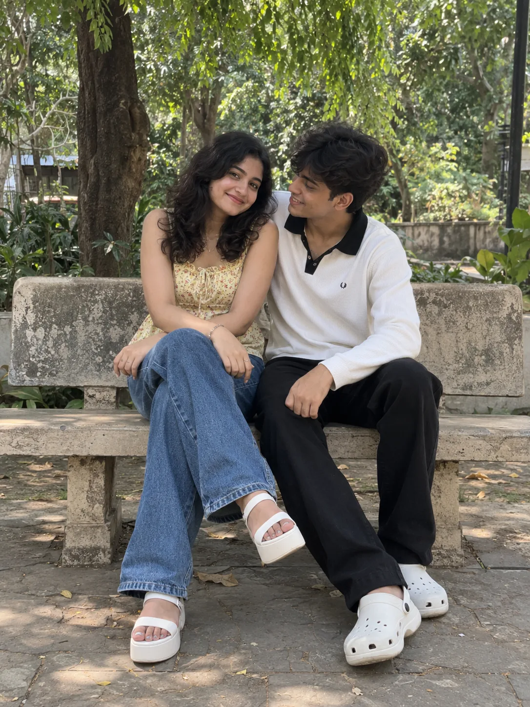 A realistic, full-body photograph of a happy young Indian couple sitting close together on a weathered conc rete park bench outdoors.