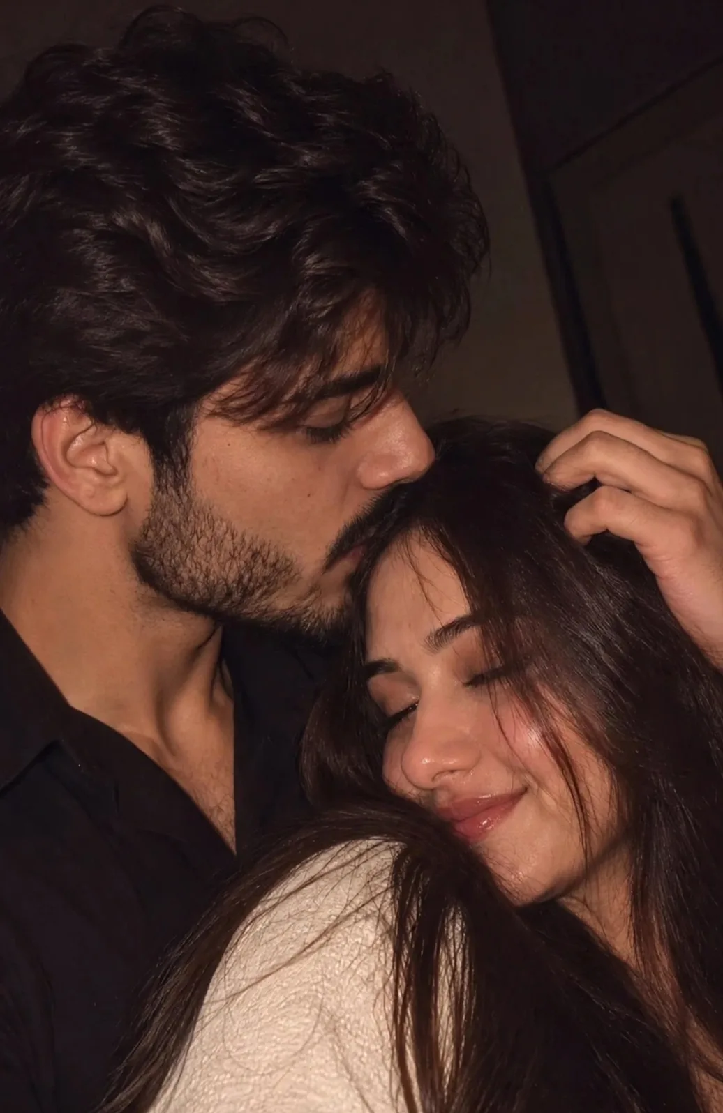 Photorealistic romantic close-up selfie of a young couple cuddling cheek-to-cheek in a dimly lit room.