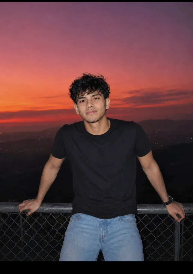 Create an ultra-realistic cinematic portrait photograph of a young man standing at a scenic hilltop viewpoint during a dramatic sunset.