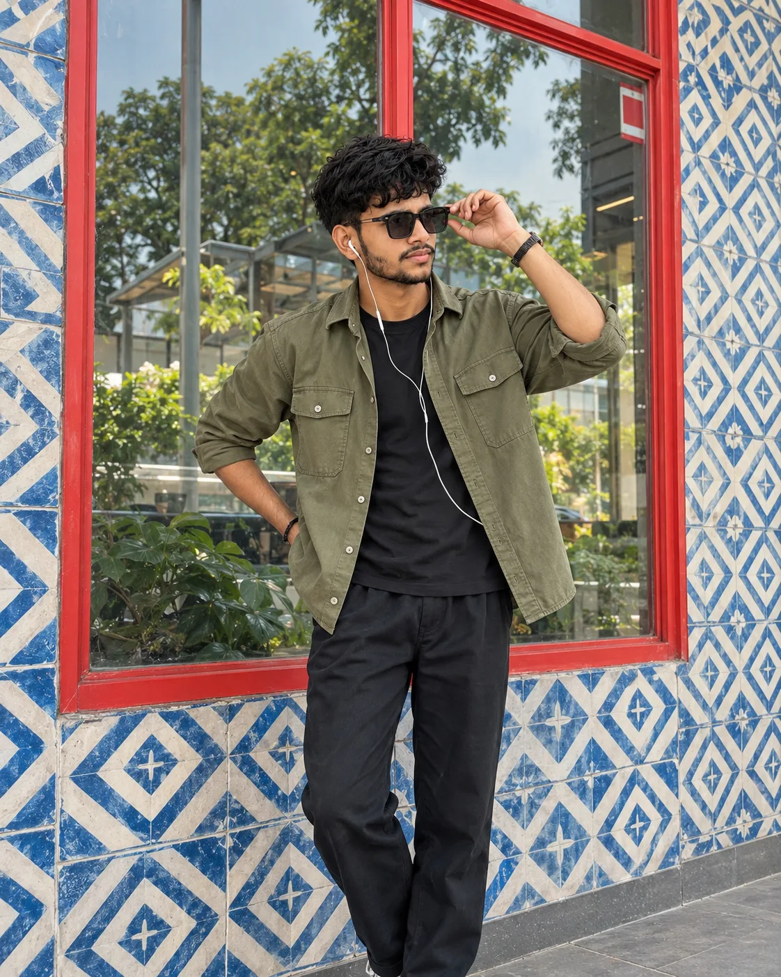 Ultra-realistic professional DSLR photograph of a young slim South Asian man standing casually in front of a large glass window framed with