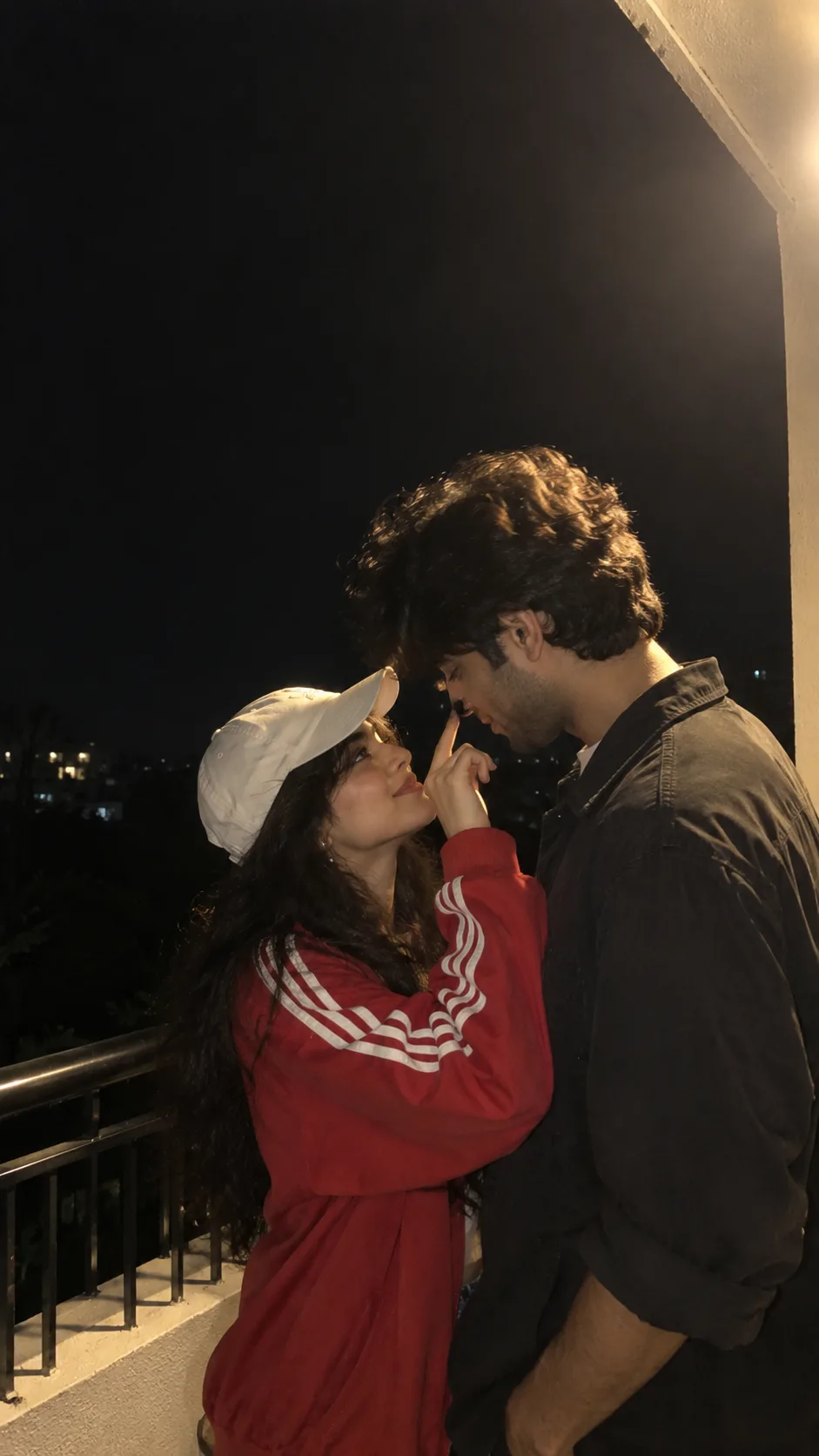 Create an ultra-realistic candid nighttime photograph of a young South Asian couple standing very close together on a balcony or terrace.