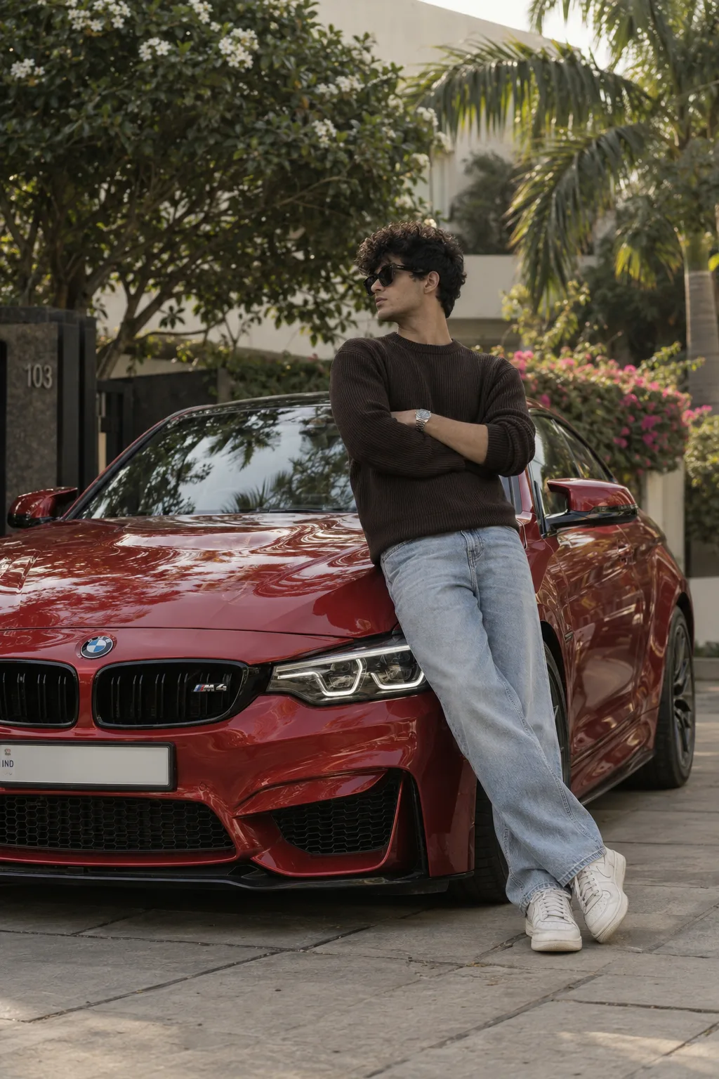 A stylish young man with curly hair, wearing black sunglasses, leaning casually against a shiny red BMW M4 in an outdoor residential