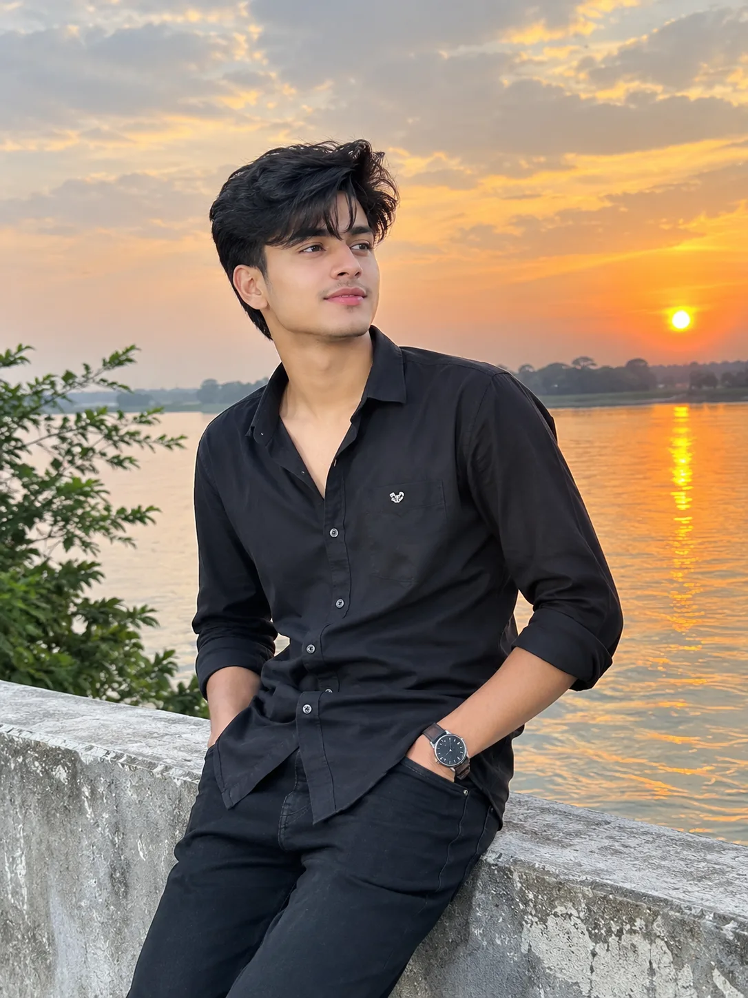 A stylish young South Asian man wearing a black shirt and black trousers, sitting beside a peaceful river during golden hour.