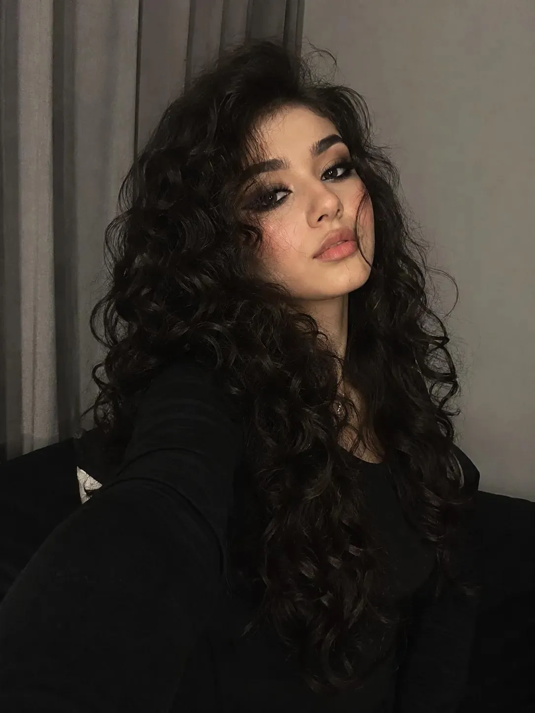 Prompt: A realistic close-up indoor selfie portrait of a beautiful young woman with very long, thick, voluminous dark black curly hair,