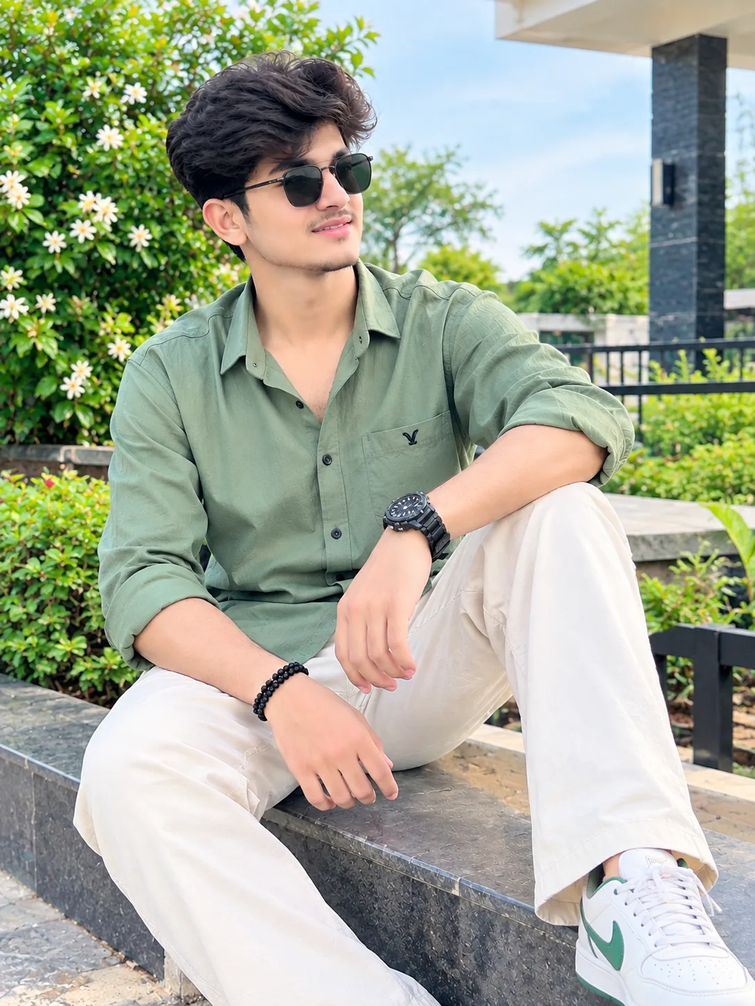 A handsome young South Asian man wearing a stylish sage green shirt, cream trousers, white sneakers, and black sunglasses, sitting