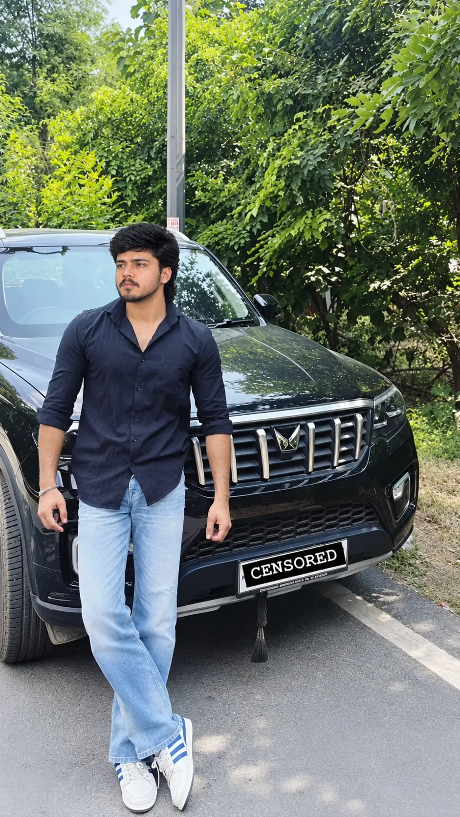 A photorealistic full-body portrait of a young South Asian man casually leaning against a glossy black luxury SUV on a leafy roadside.
