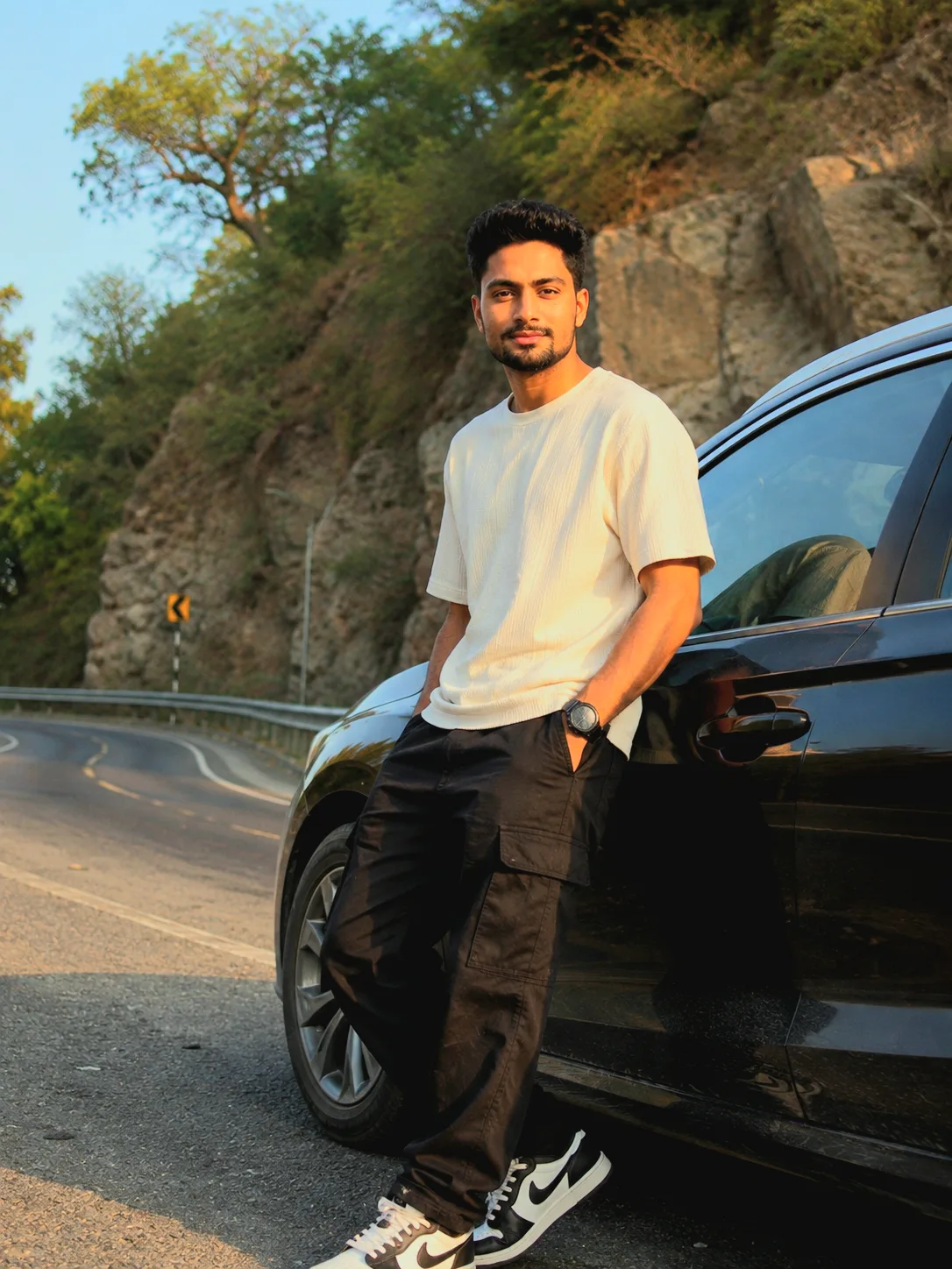 Create an ultra-realistic DSLR portrait of me casually leaning against the front passenger side of a glossy black sedan on a scenic