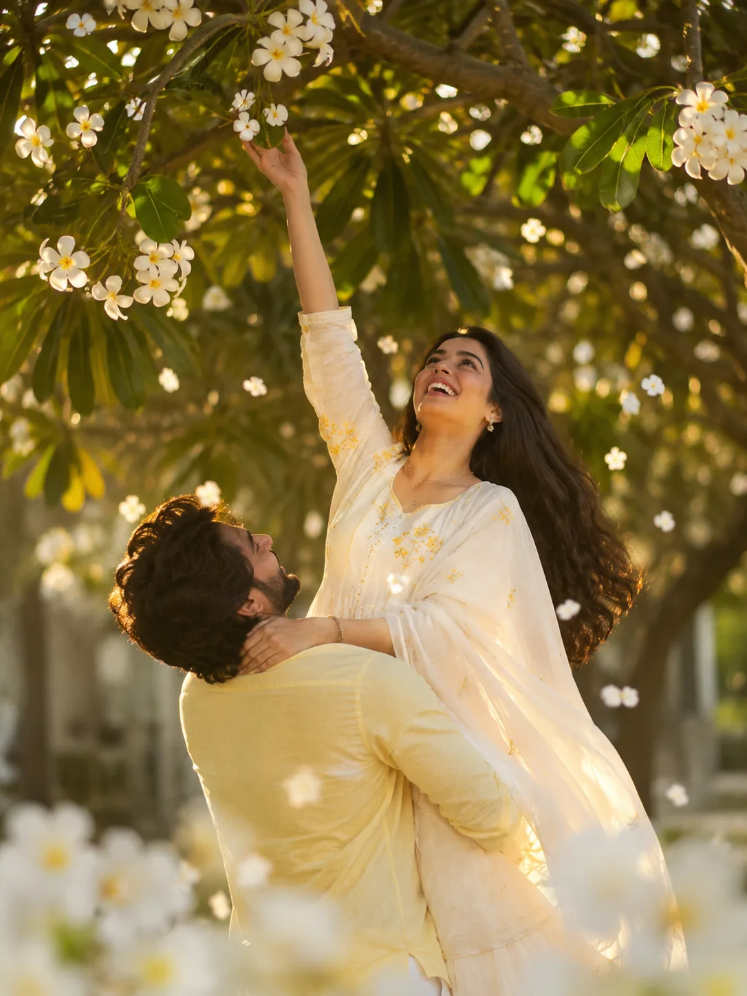A candid, romantic medium shot of a young South Asian couple under a lush Frangipani (Plumeria) tree during golden hour.