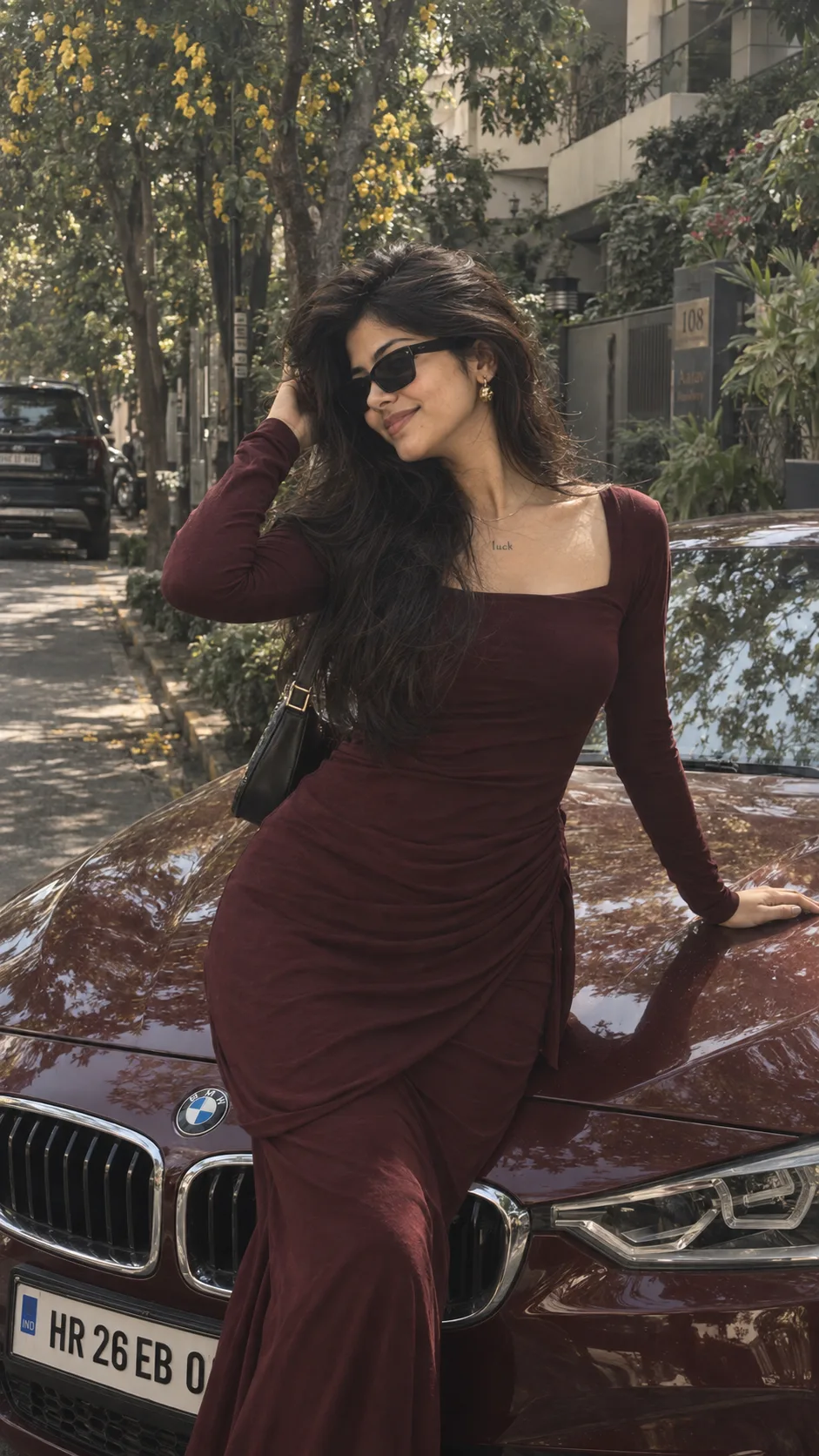 A stylish, realistic outdoor portrait of a beautiful young Indian woman standing and leaning against the hood of a maroon BMW car,