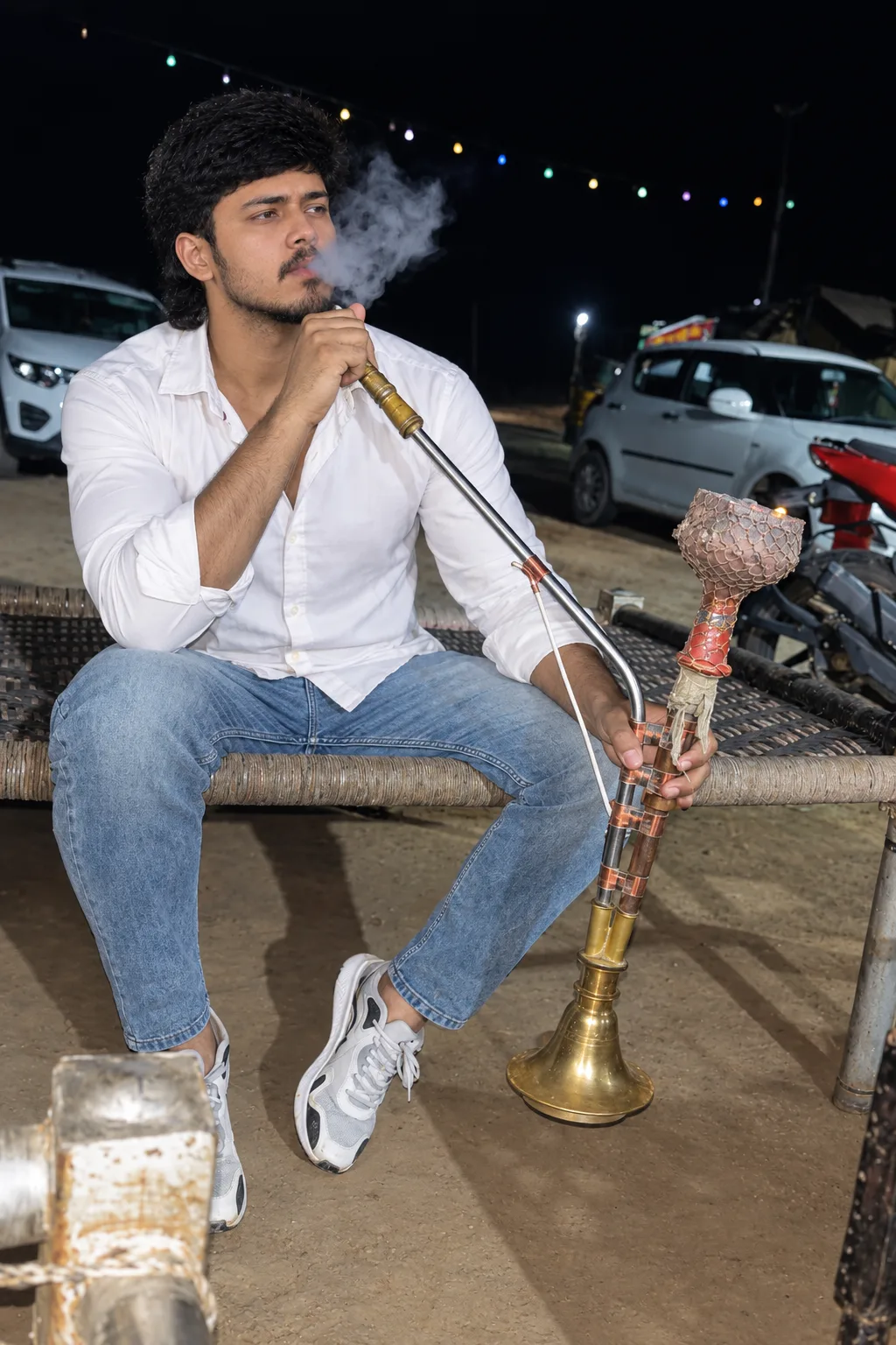 Create a highly realistic nighttime lifestyle photograph of a young South Asian man sitting casually on a traditional woven charpoy,