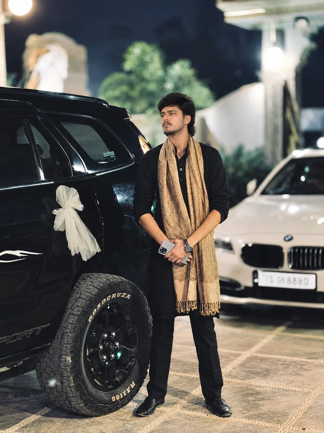 Create a photorealistic cinematic full-body portrait of a young Indian man standing confidently outdoors at night beside a luxurious black