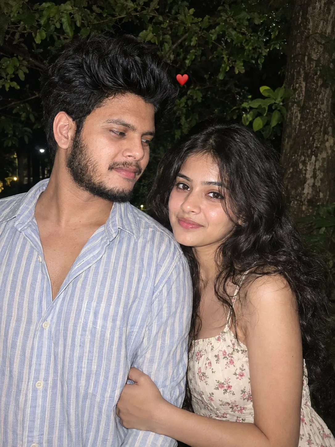 Intimate candid nighttime portrait of a young South Asian couple standing close outdoors against a dark leafy background, waist-up