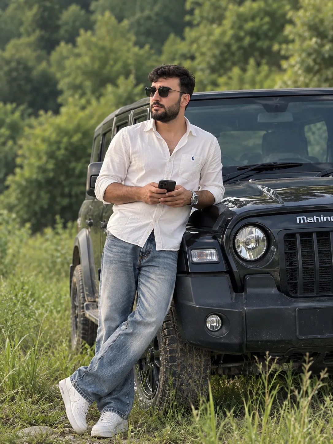 A cinematic outdoor portrait of a stylish young South Asian man with wavy dark hair and a short beard, wearing dark sunglasses, a