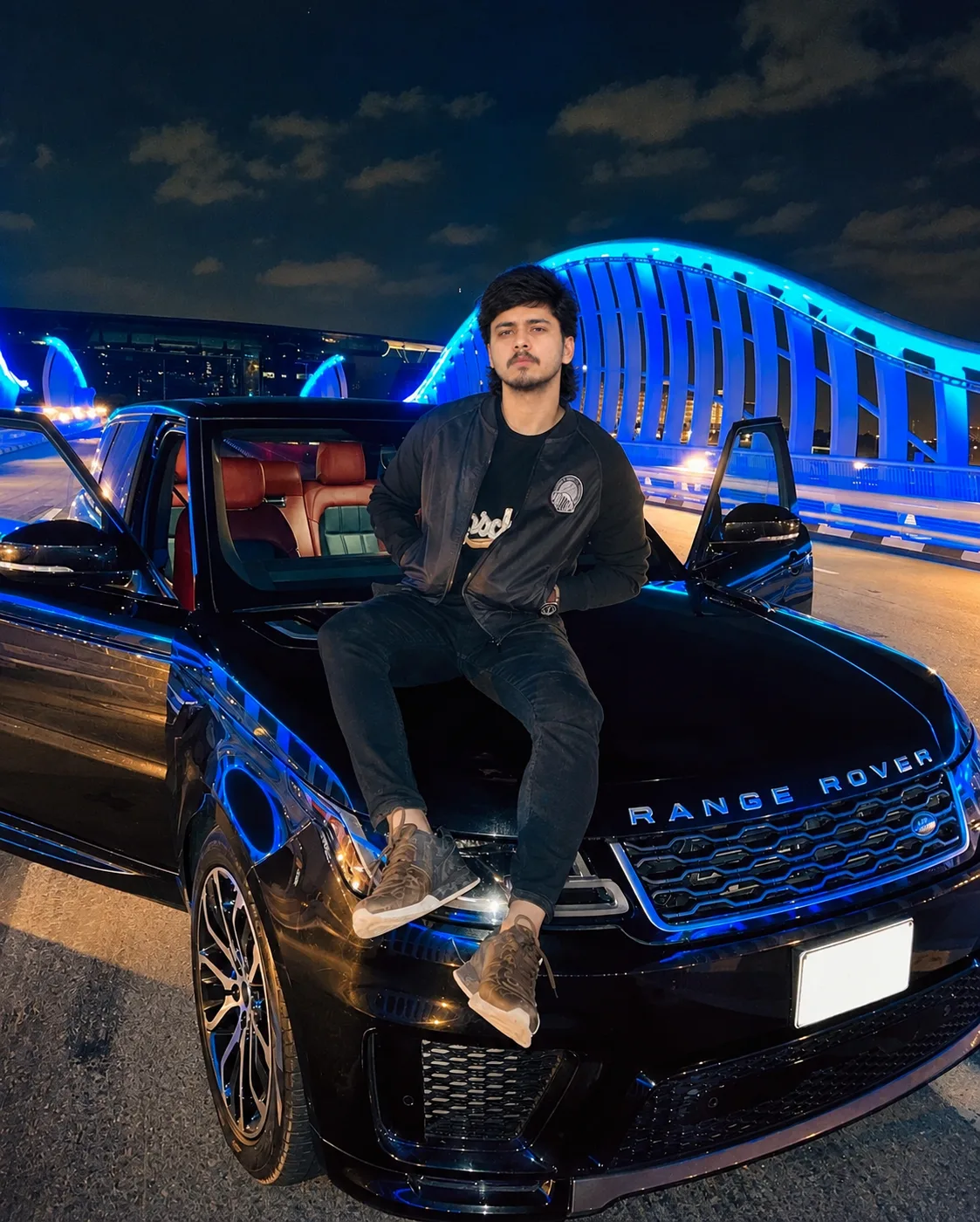 Create an ultra-realistic, cinematic vertical night portrait of a young South Asian/Indian man sitting confidently on the front hood of a