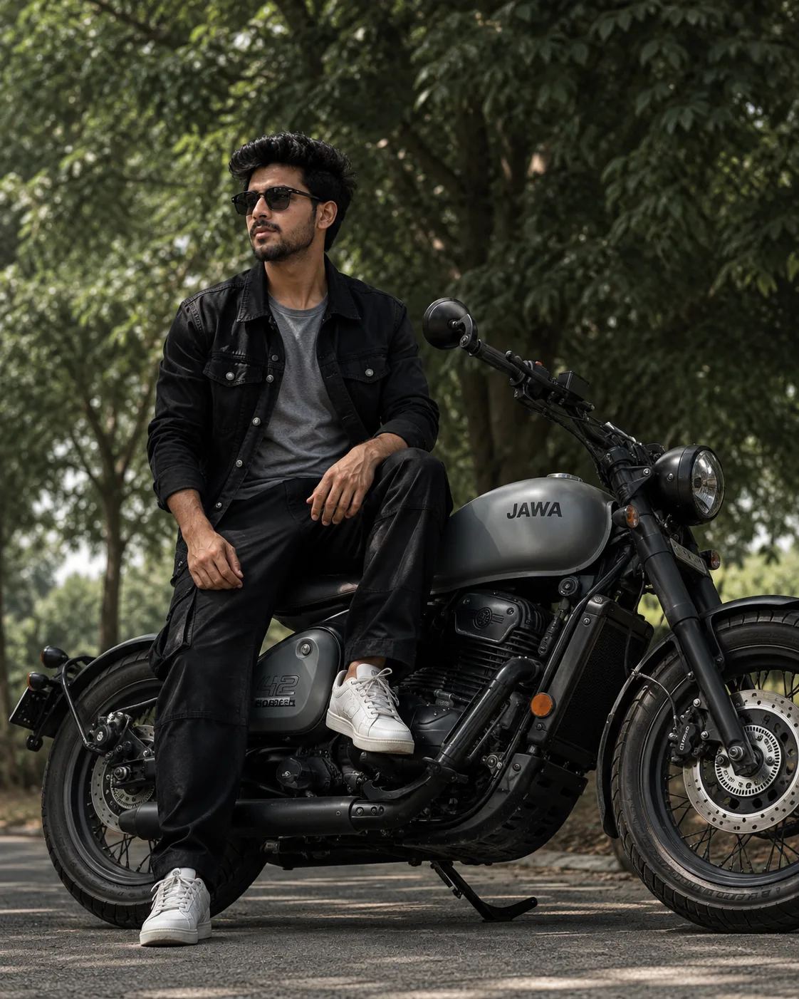 Ultra-realistic full-body shot of a stylish young man sitting confidently on a matte grey Jawa 42 bobber motorcycle under lush green trees.
