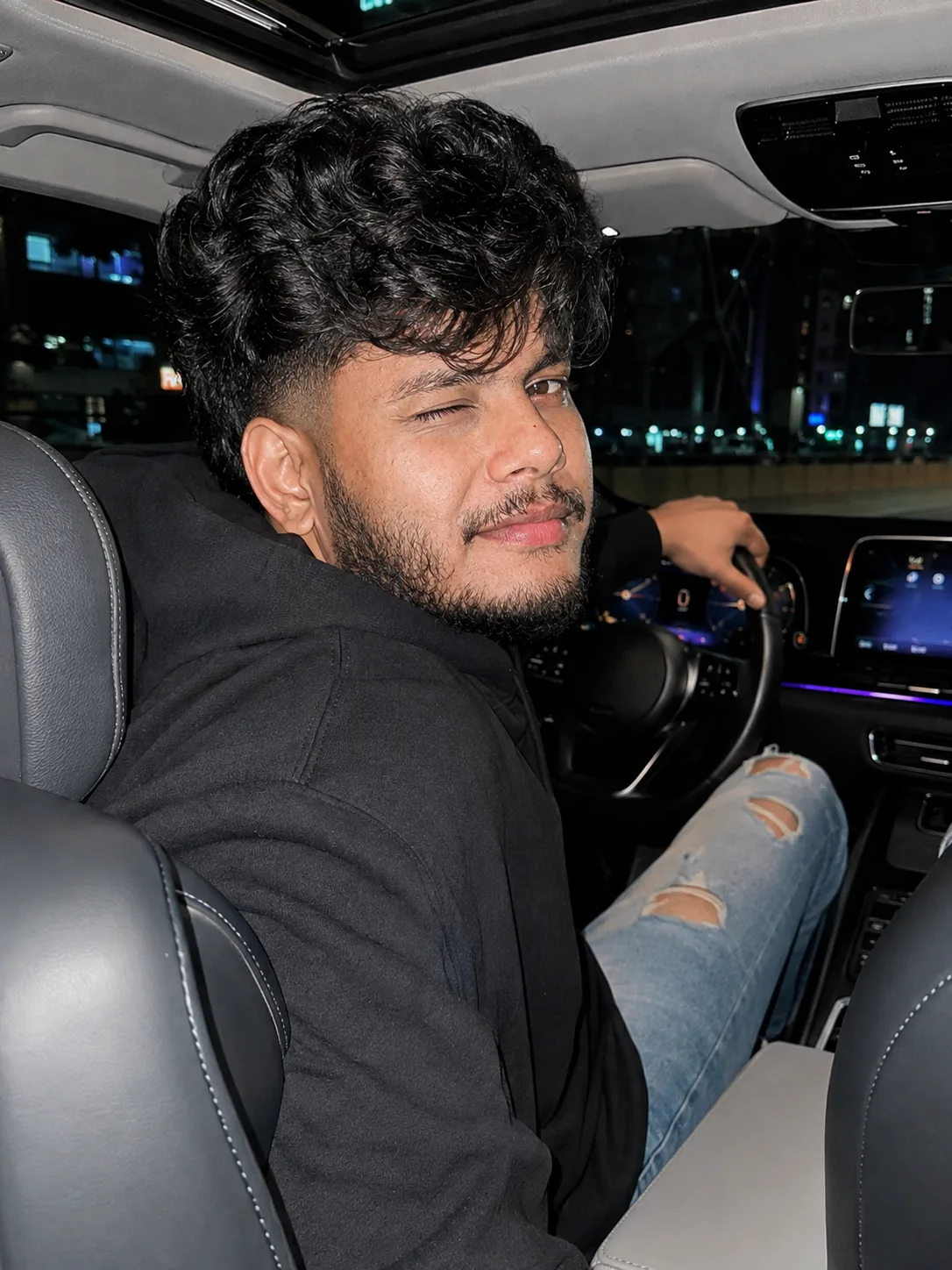 A young Indian man in his early 20s sitting in the driver’s seat of a modern car at night, turning his body slightly toward the camera.