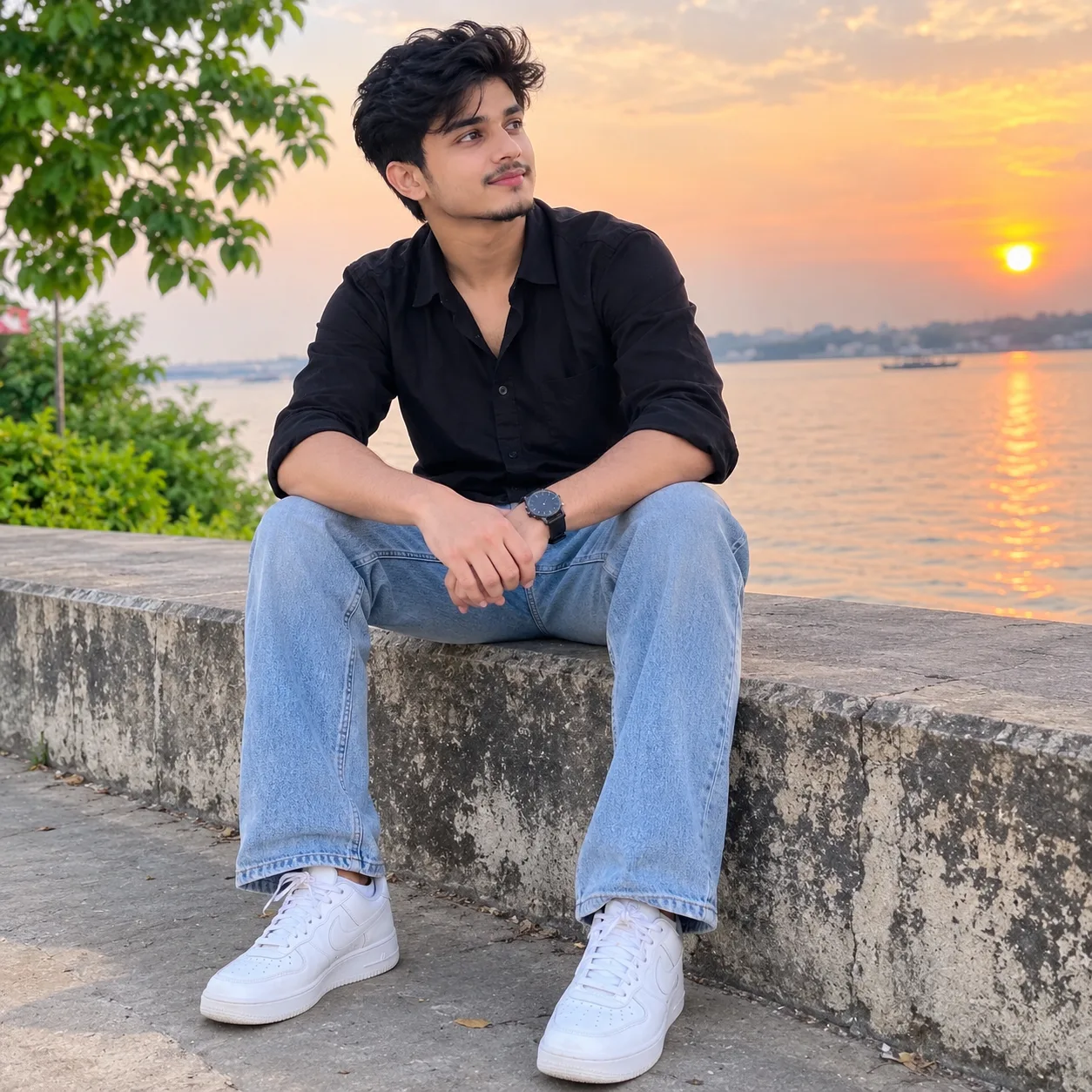 A handsome young South Asian man sitting naturally beside a beautiful riverside during golden hour, full body clearly visible, stylish
