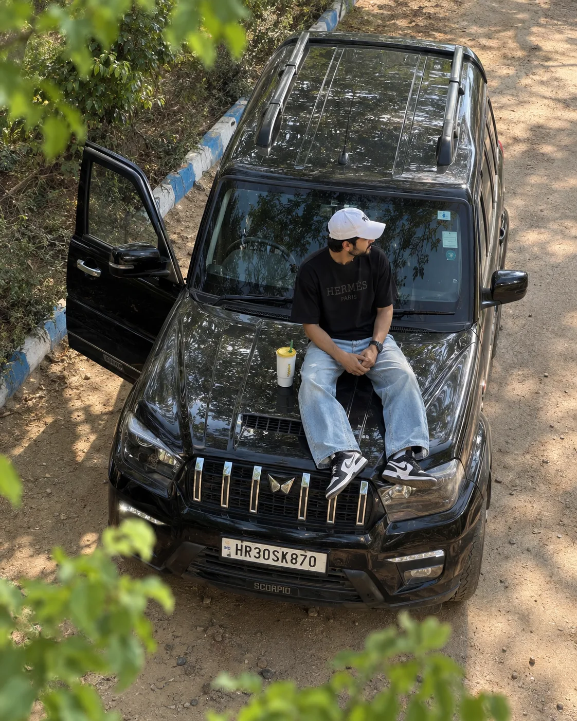 @Create image High-angle aerial top-down drone shot of a young Indian man in his mid-20s with a light beard sitting on the bonnet / hood of