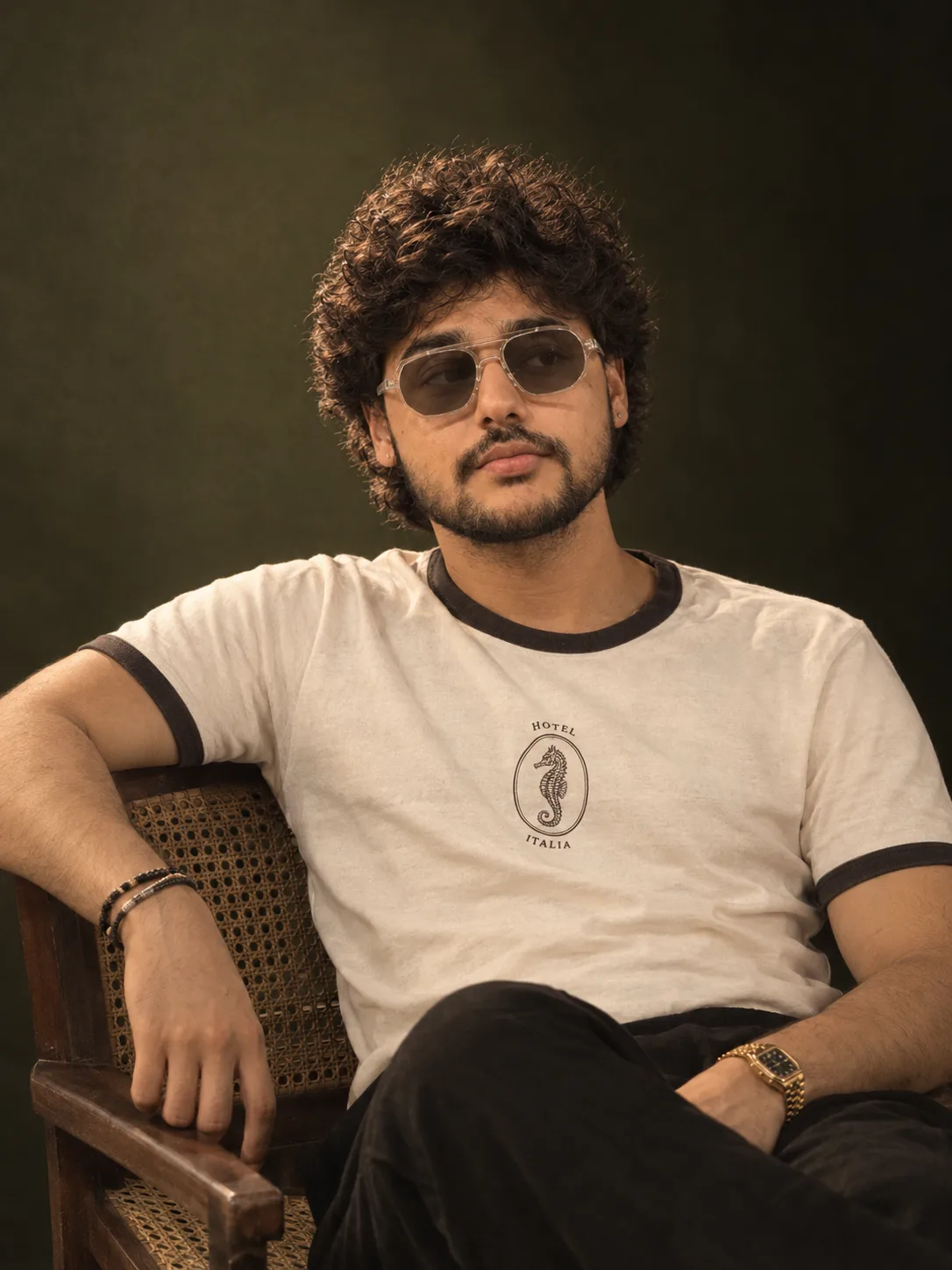 "A polished studio portrait of a young man with thick, soft, tousled black wavy hair, light stubble, and a trimmed mustache, gazing off to