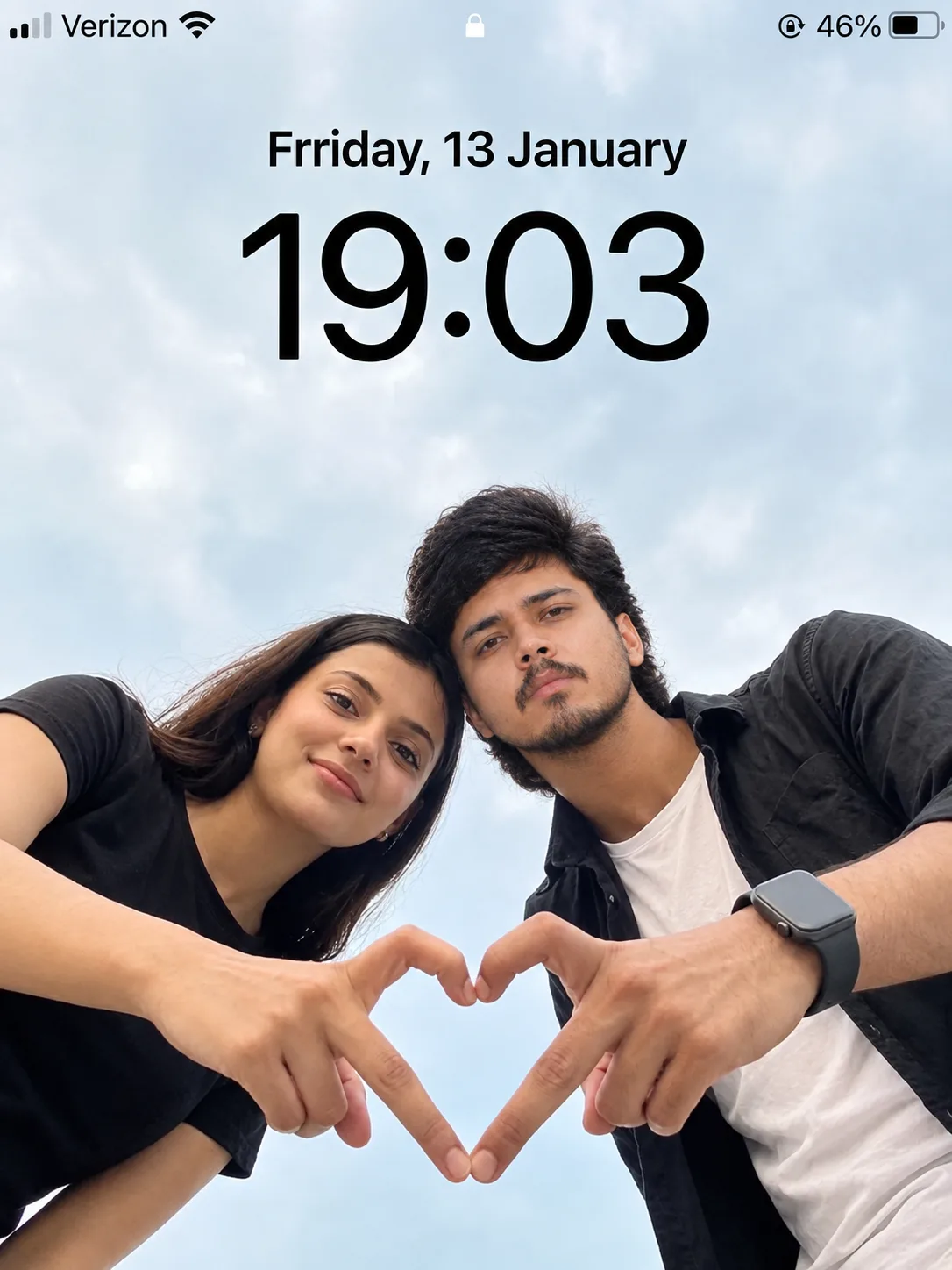 A photorealistic low-angle outdoor portrait of a young South Asian couple leaning toward the camera against a bright blue sky filled with