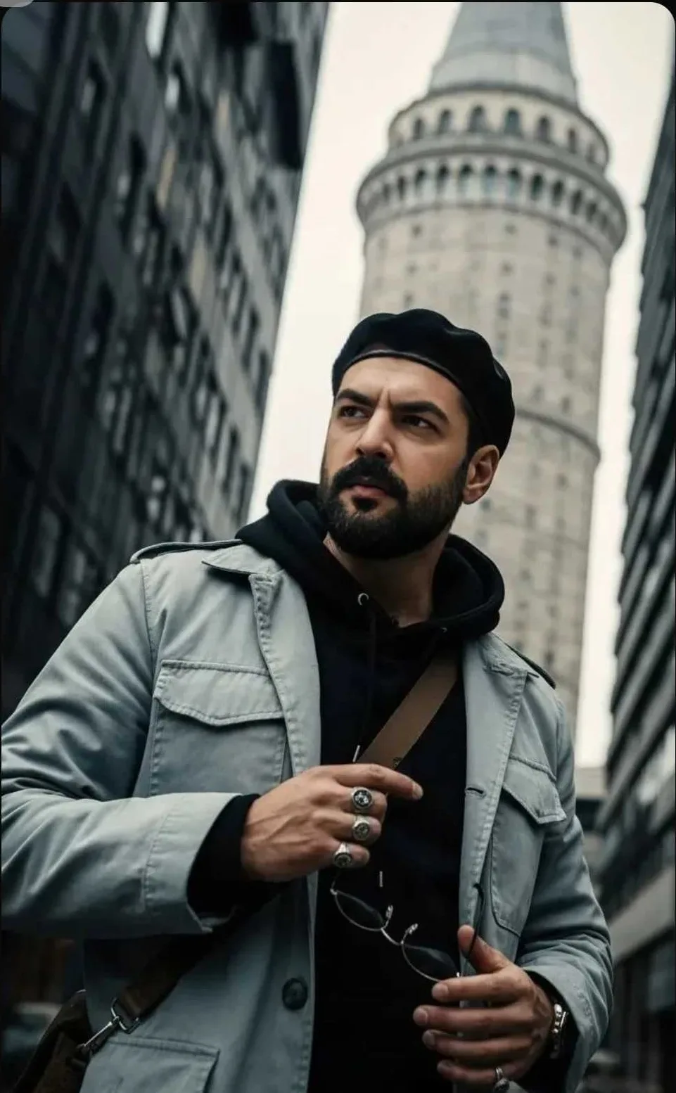 Cinematic portrait of a bearded man wearing black beret, black hoodie and light grey jacket, holding sunglasses, standing in old city