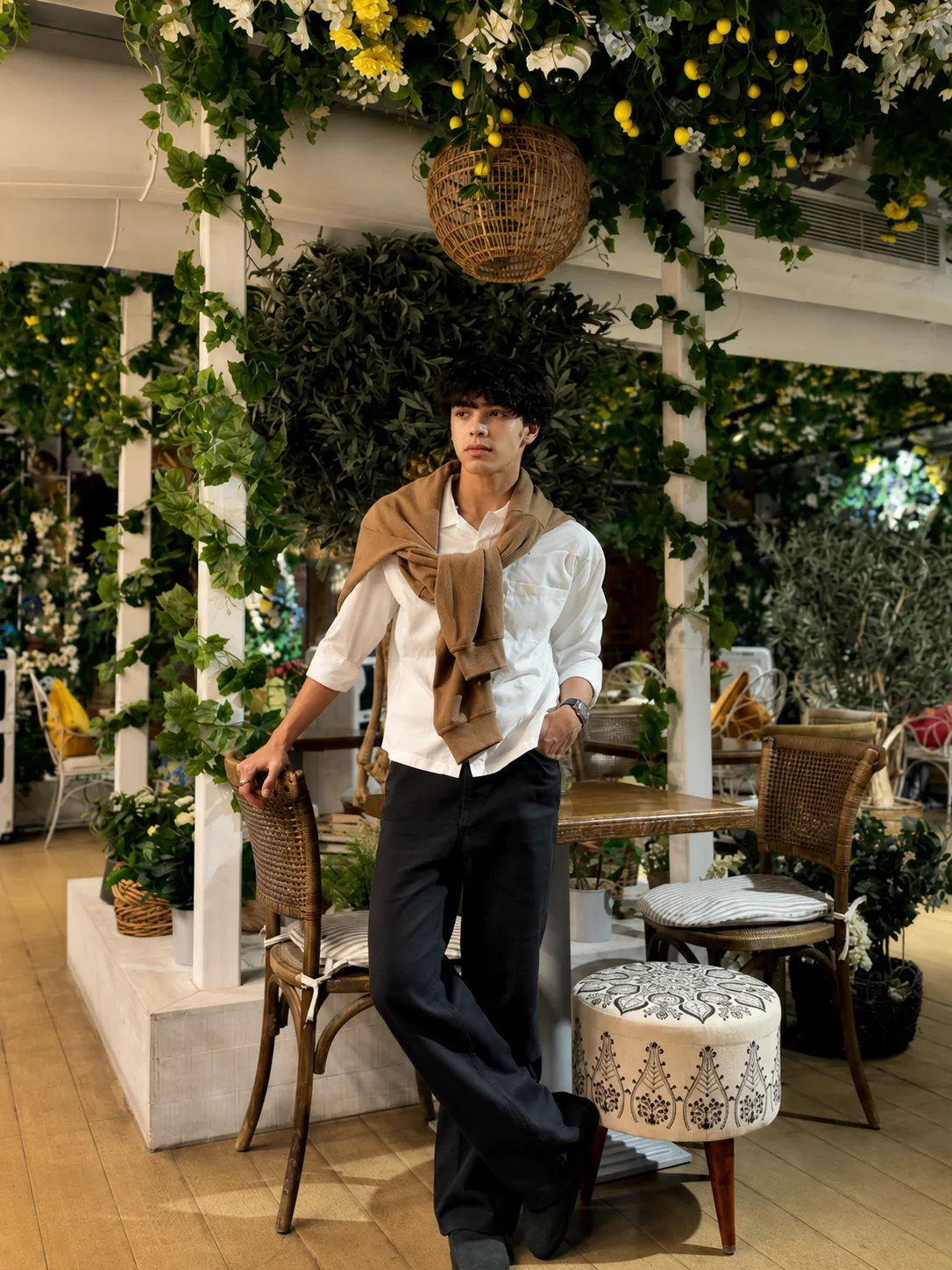 An exceptionally stylish young male fashion model standing confidently inside an elegant luxury botanical café filled with lush greenery,