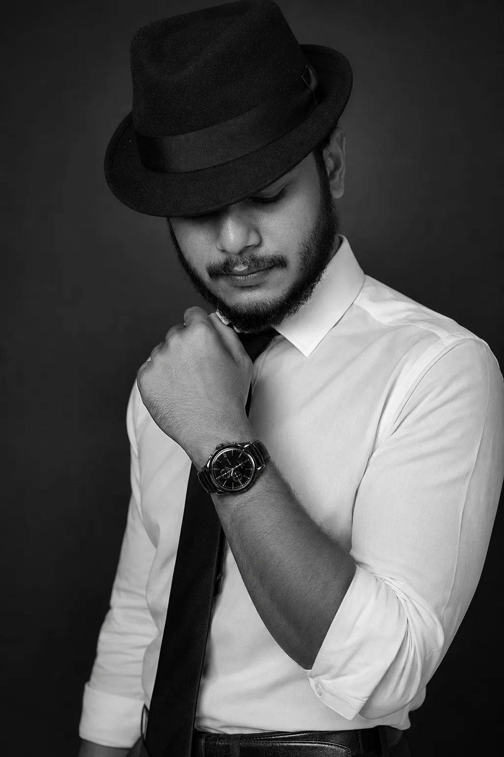 Black and white studio portrait of a stylish young Indian man with a neatly trimmed full beard and mustache, wearing a classic black fedora