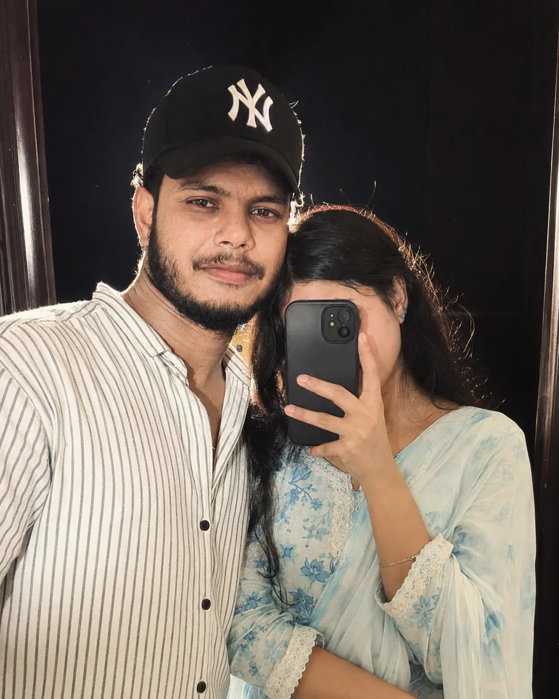 A realistic mirror selfie of a young Indian couple standing close together indoors against a dark background.
