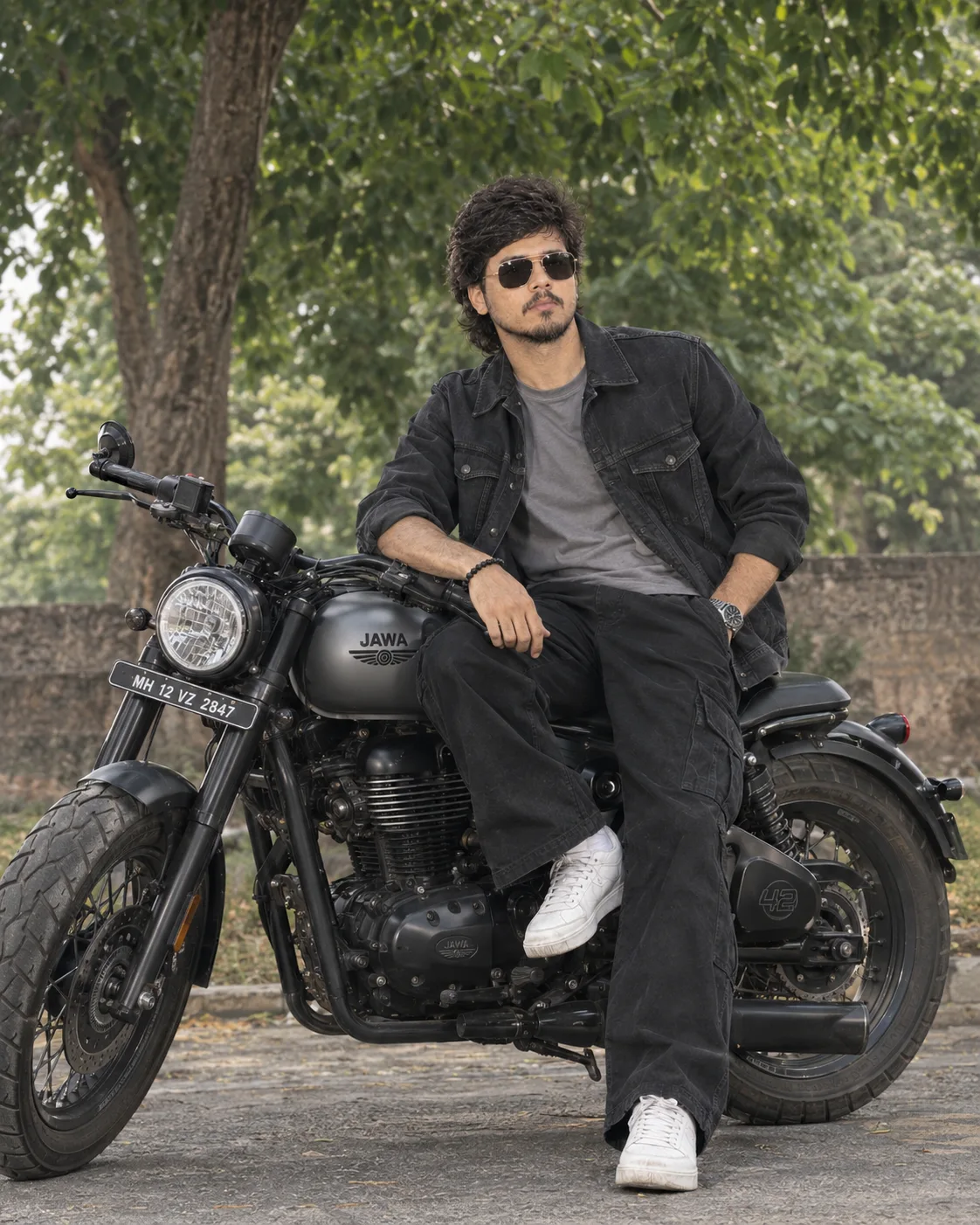Ultra-realistic full-body shot of a stylish young man sitting confidently on a matte grey Jawa 42 bobber motorcycle under lush green trees.