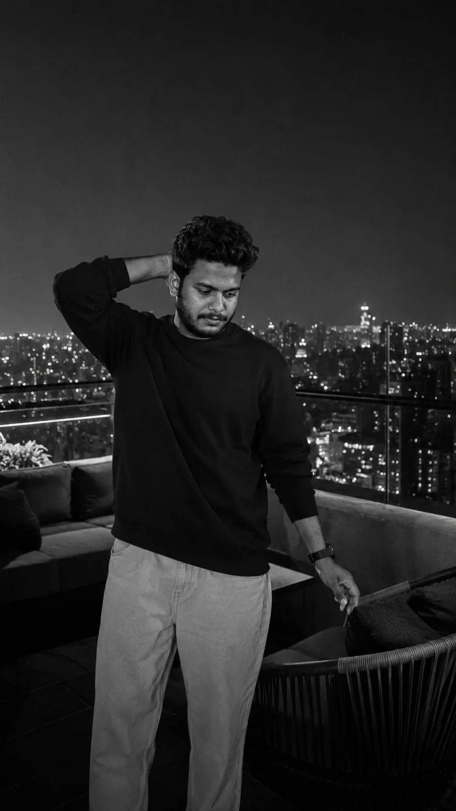 Black and white cinematic portrait of a young Indian man with curly dark hair and light beard standing on a modern rooftop terrace at