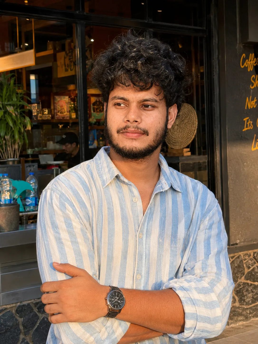 A ultra-realistic cinematic portrait of a 26-year-old Indian man standing outdoors near a modern cafe storefront during golden hour.