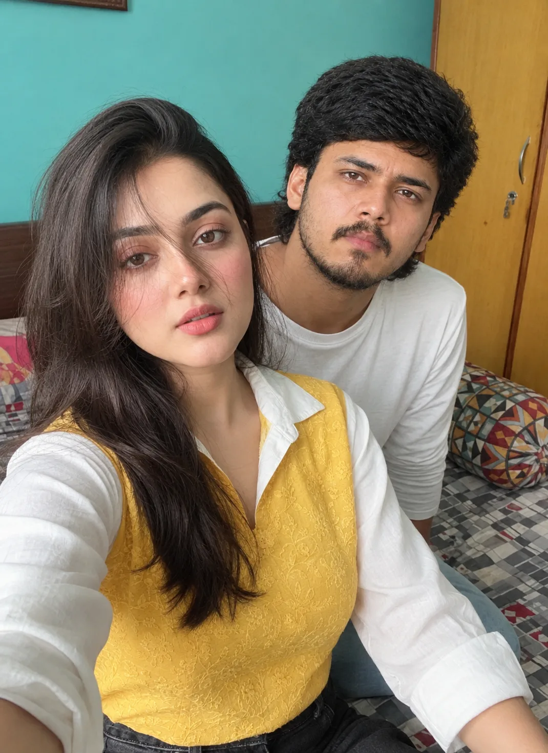 A natural, ultra-realistic indoor selfie of a young couple sitting closely together on a bed in a cozy bedroom.