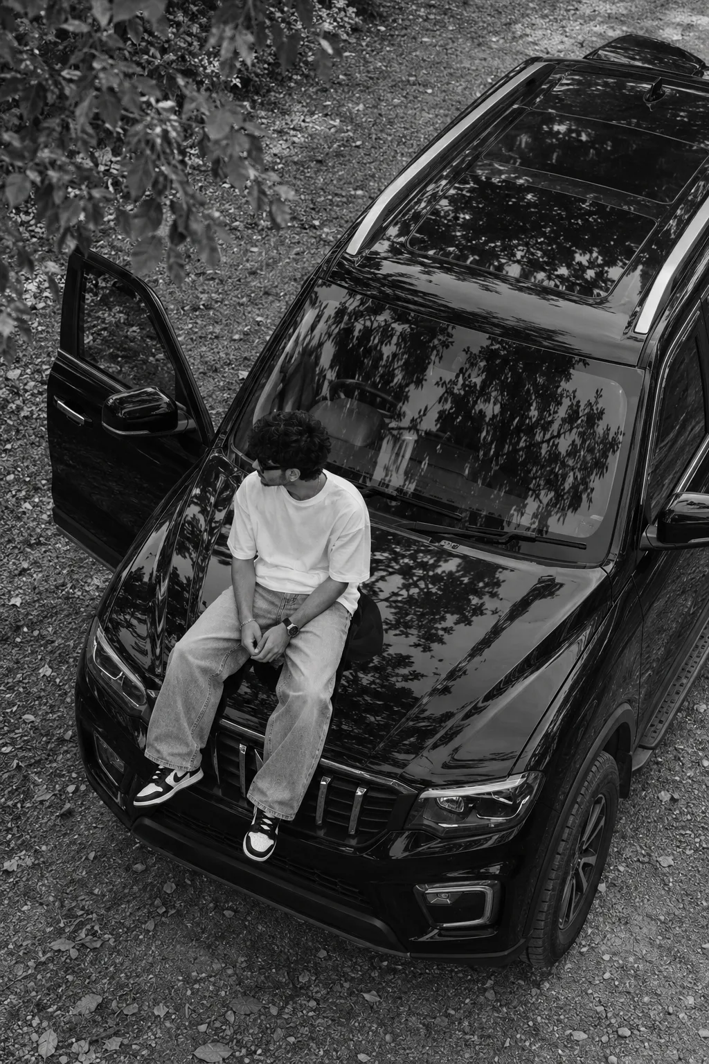 Create a highly realistic, cinematic, black and white photograph of a stylish young person sitting on the hood of a black SUV, captured