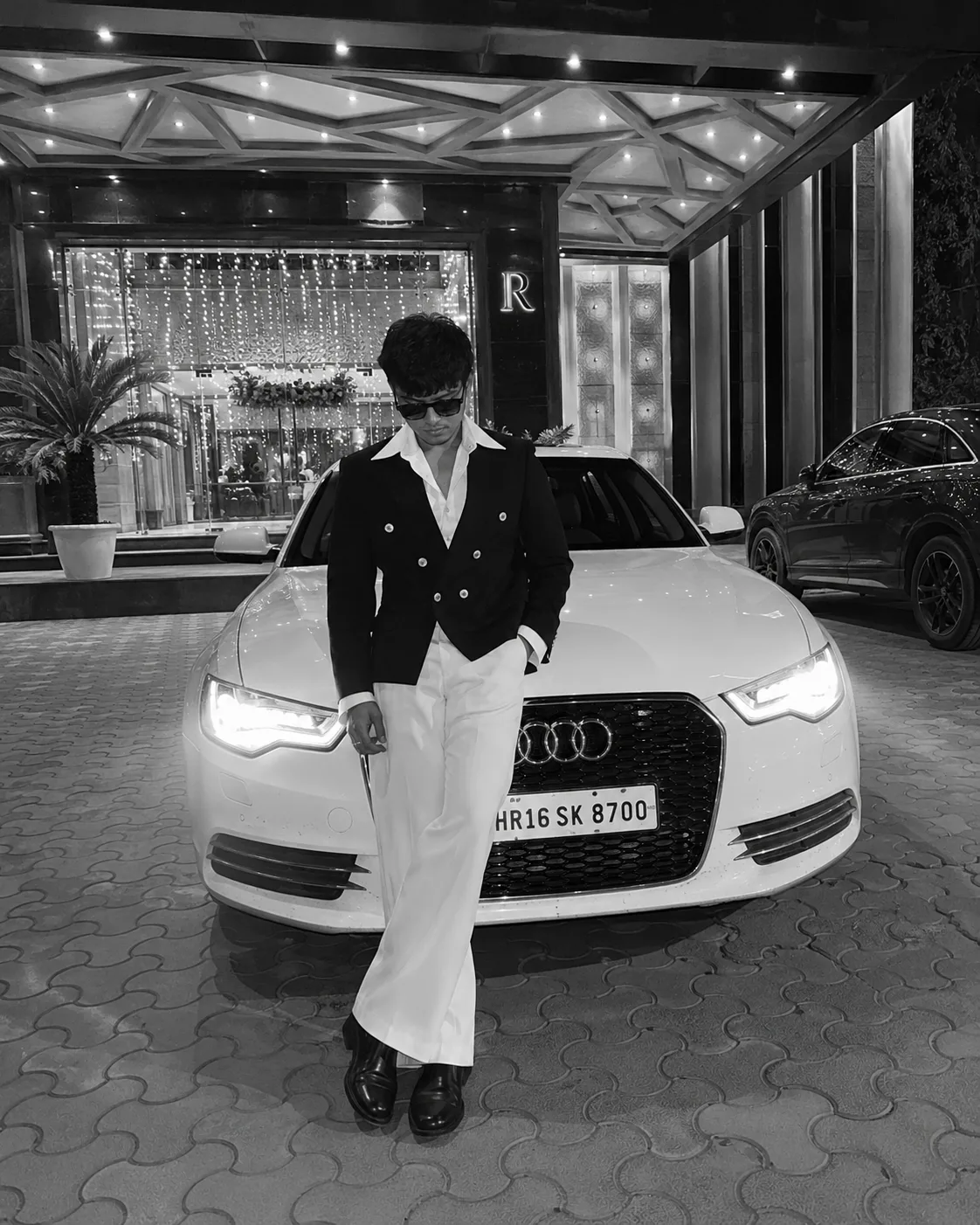 Black and white picture Photorealistic full-body night photo of a young South Asian stylish man in his mid-20s leaning casually against the
