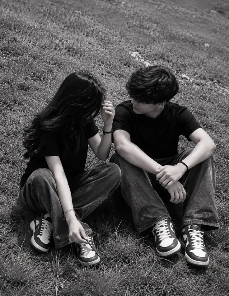 A high-contrast black and white candid photograph of a young couple sitting cross-legged on a grassy slope, facing each other.
